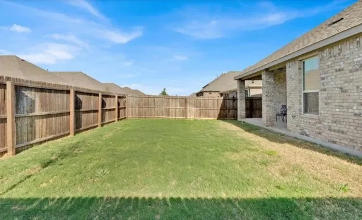 Expansive backyard featuring a green lawn, wood privacy fencing, a covered patio, and a brick exterior
