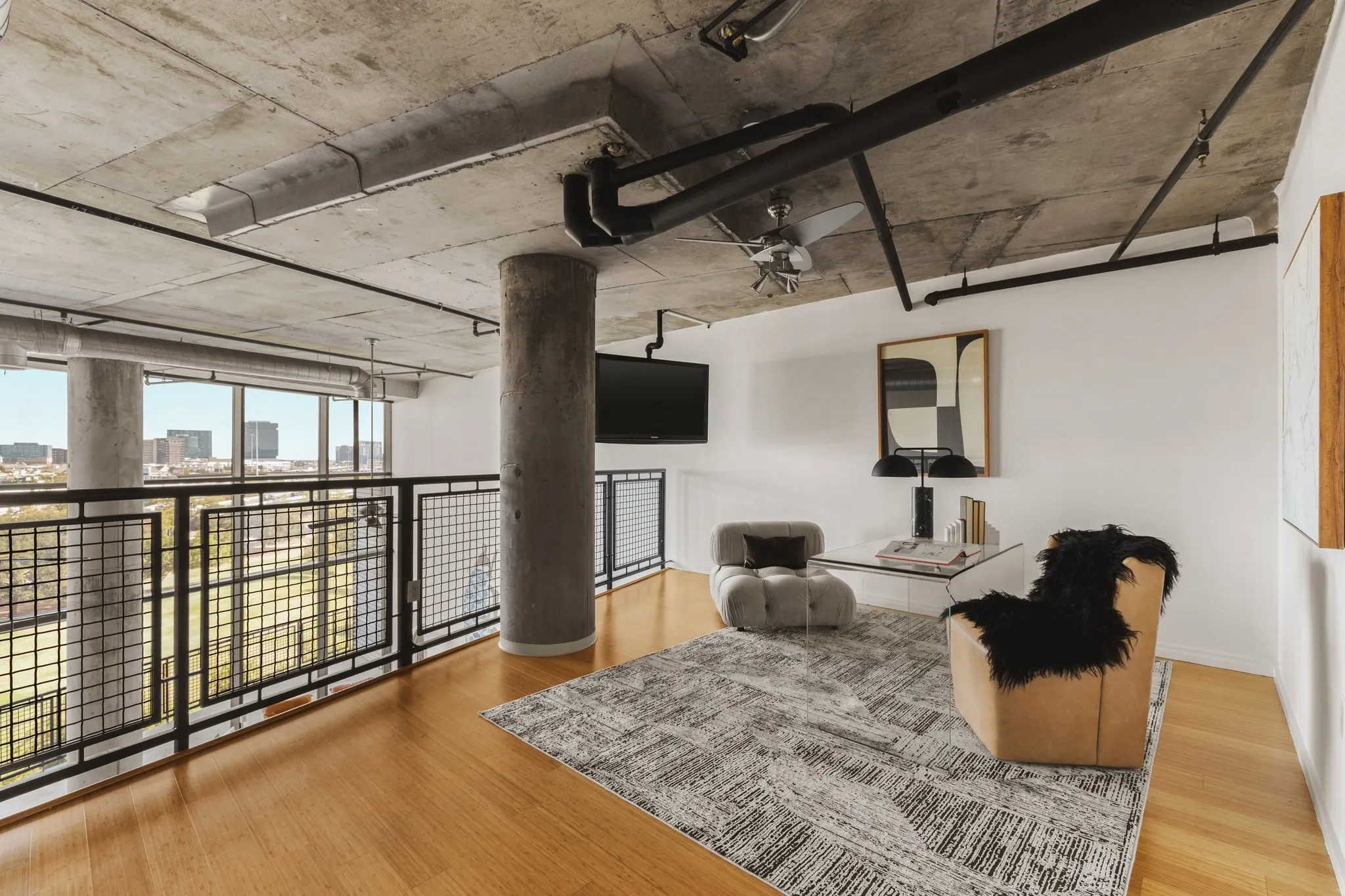 Exposed concrete ceiling with industrial piping, wood-finish flooring, full-height windows, and metal grid balcony railing
