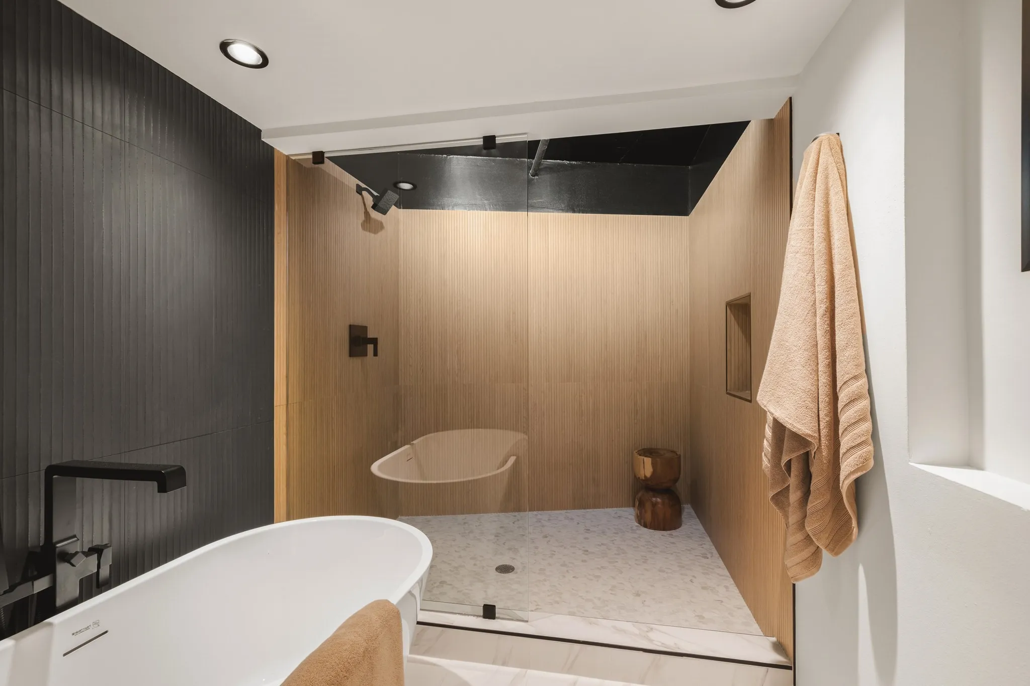 Freestanding soaking tub with a wall-mounted matte black faucet, walk-in shower with light wood-finish walls, glass enclosure, and recessed lighting