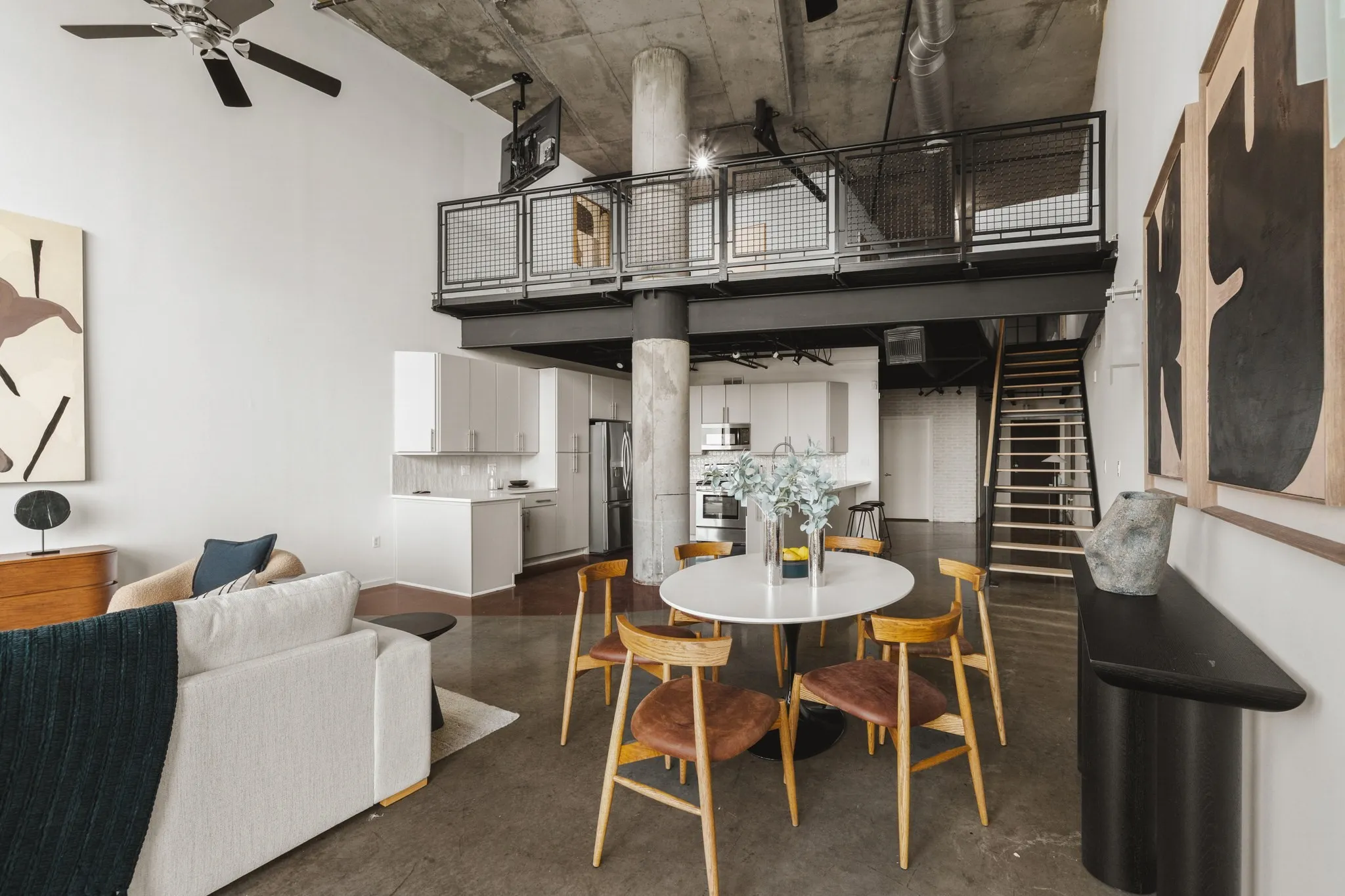 High ceilings with exposed concrete, polished concrete flooring, open-concept kitchen with white cabinetry, and an upper-level mezzanine with metal railing