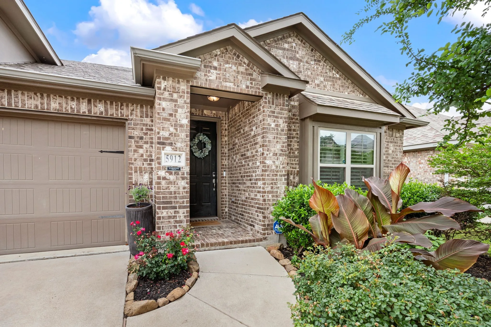 Charming brick home featuring a welcoming front entrance with black accent door, dual-pane windows, and professional landscaping including flowering beds and lush greenery