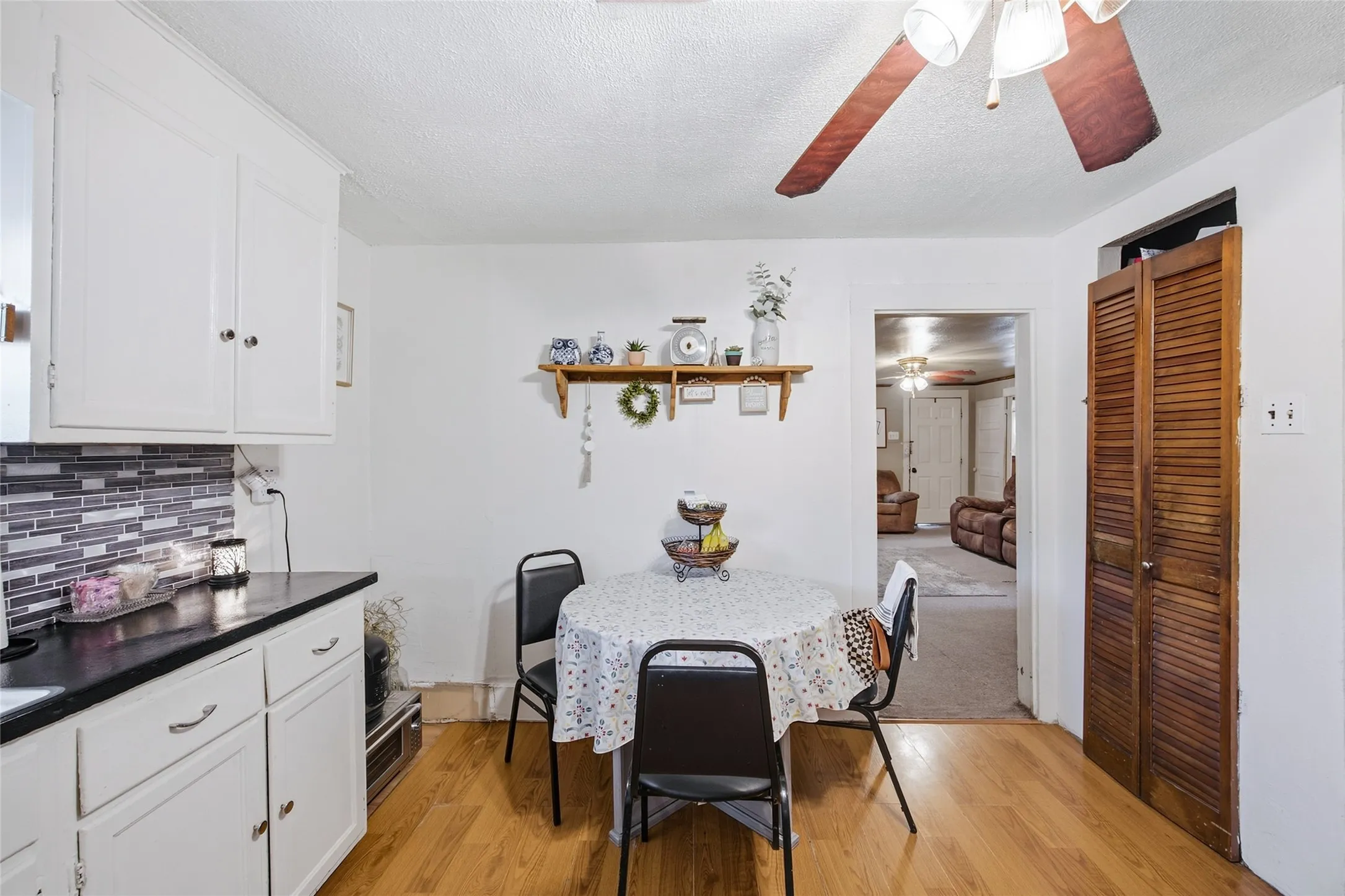 White cabinetry with dark countertops, a mosaic tile backsplash, wood-finish flooring, and a ceiling fan with integrated lighting