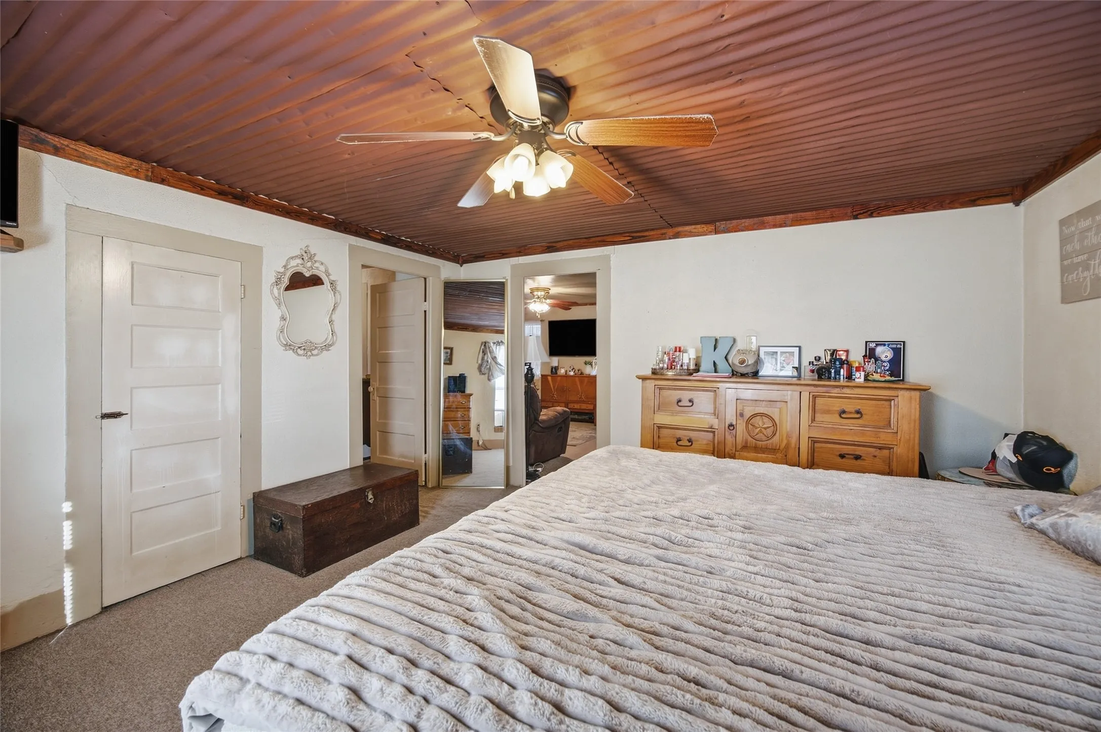 Corrugated metal ceiling with exposed wood beams, ceiling fan with light fixture, light-colored walls, and carpet flooring