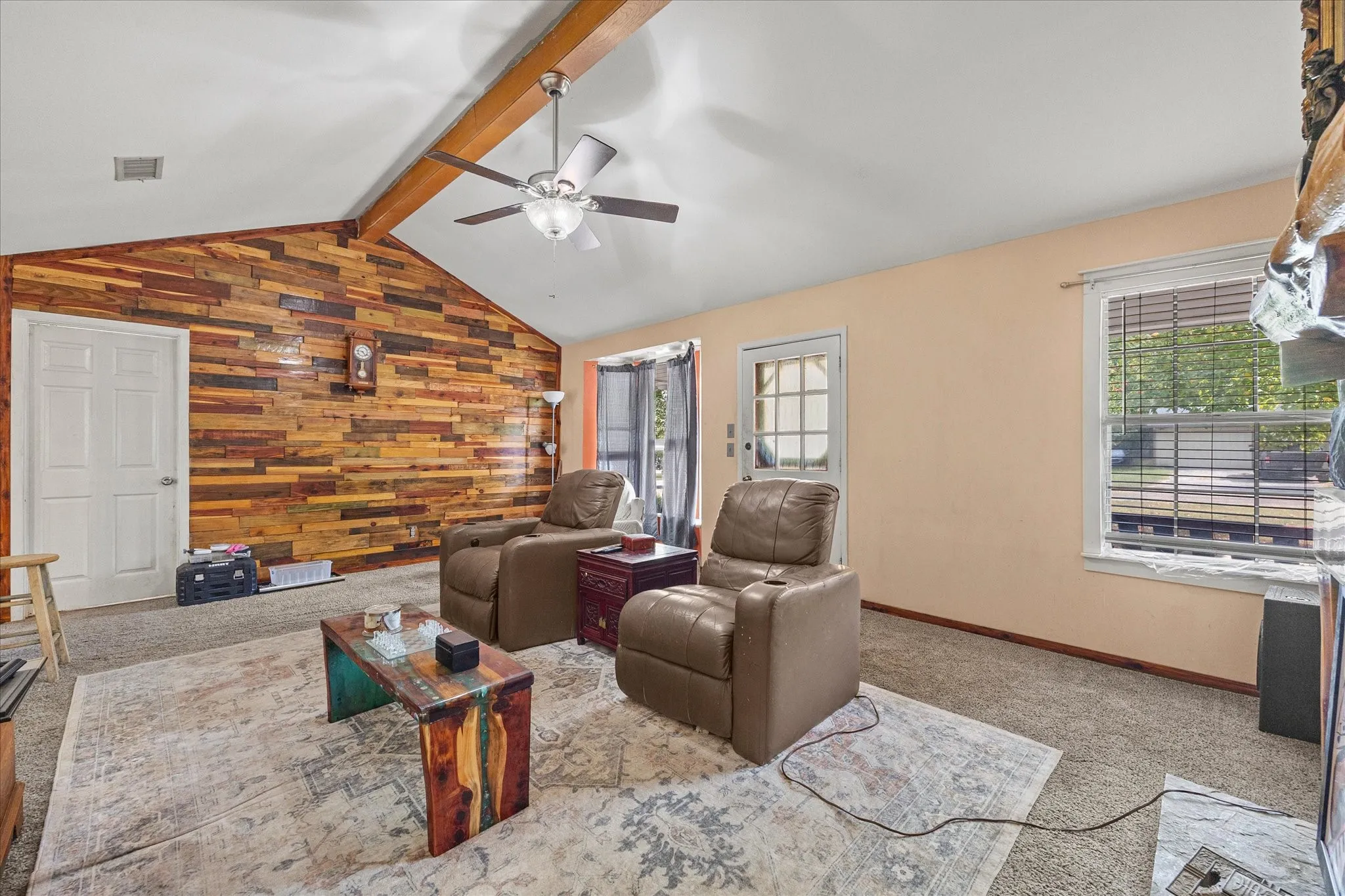Vaulted ceiling with exposed wood beam, wood plank accent wall, ceiling fan with integrated lighting, and multiple windows