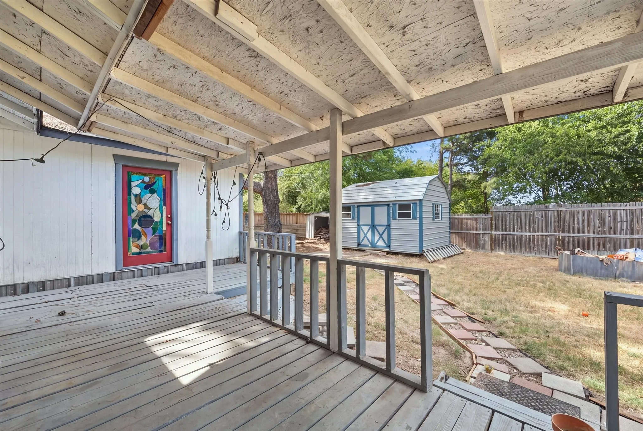 Covered outdoor deck with a decorative red door, a detached storage shed, a wood fence, and a stone-paver pathway