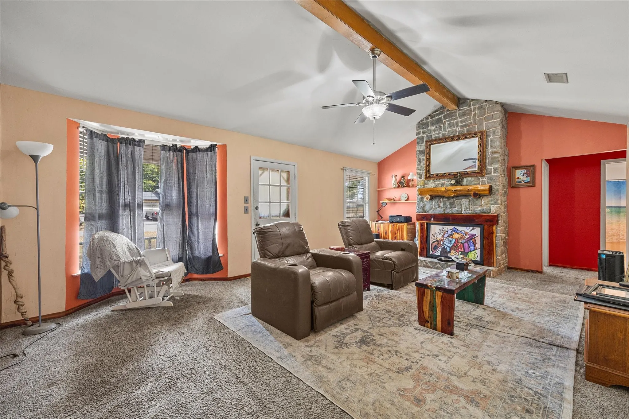 Vaulted ceilings with exposed wood beam, stone-faced fireplace with wood mantel, ceiling fan with integrated lighting, and a glass-paned exterior door