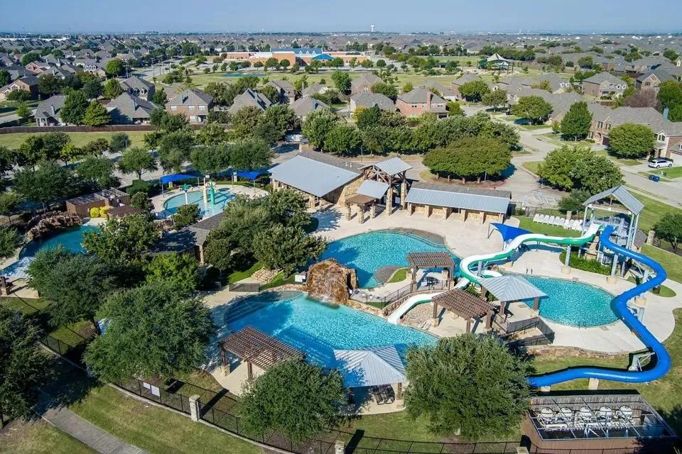 Expansive community pool area featuring multiple swimming pools, water slides, shaded lounging areas, and a central clubhouse with stone and metal roof accents