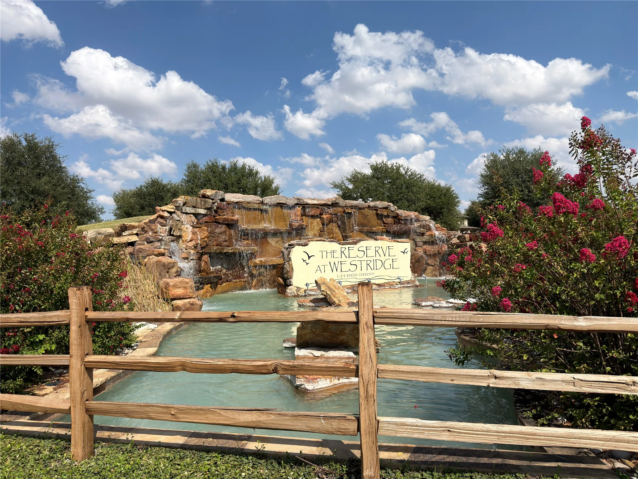 Stone waterfall feature with cascading water, surrounded by lush landscaping and blooming shrubs, with a rustic wood fence in the foreground