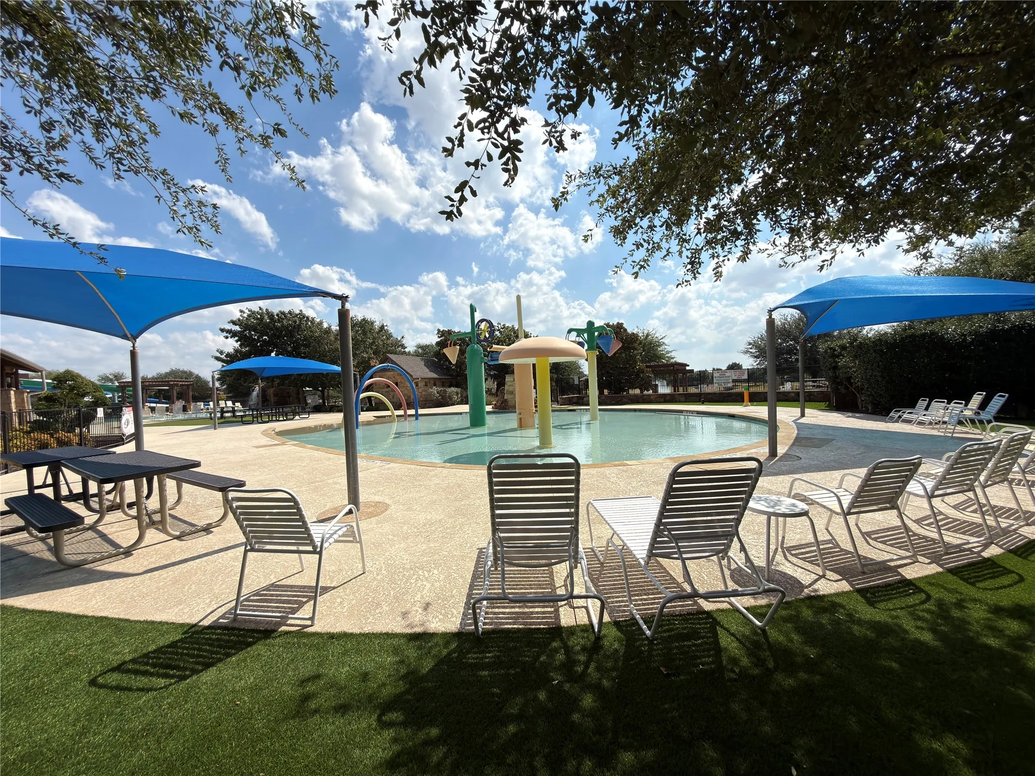 Community splash pad with multiple water features, surrounding paved deck, blue shade canopies, and artificial turf areas