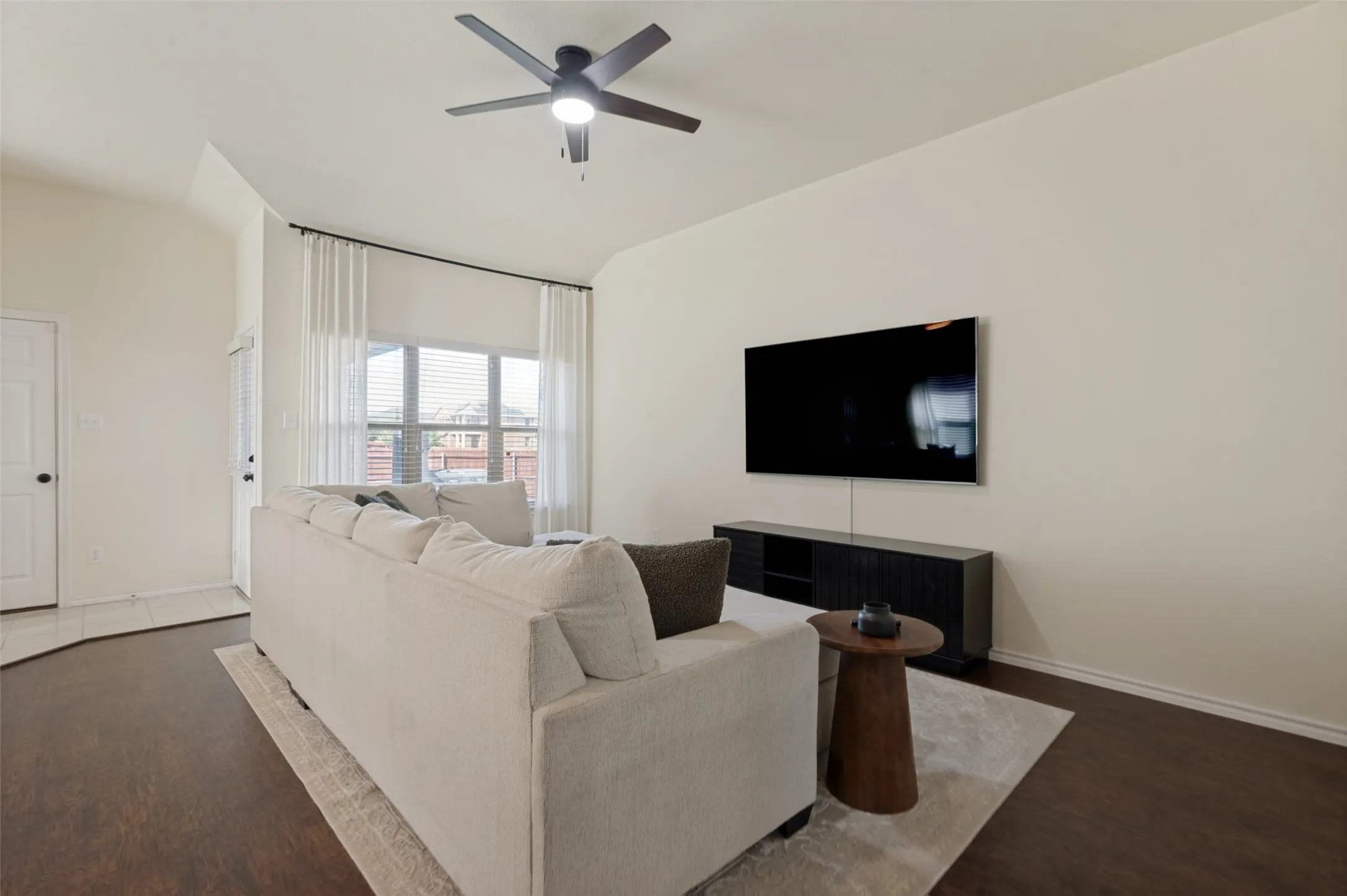Vaulted ceilings, wood-finish flooring, ceiling fan, large window with blinds, and white interior paint