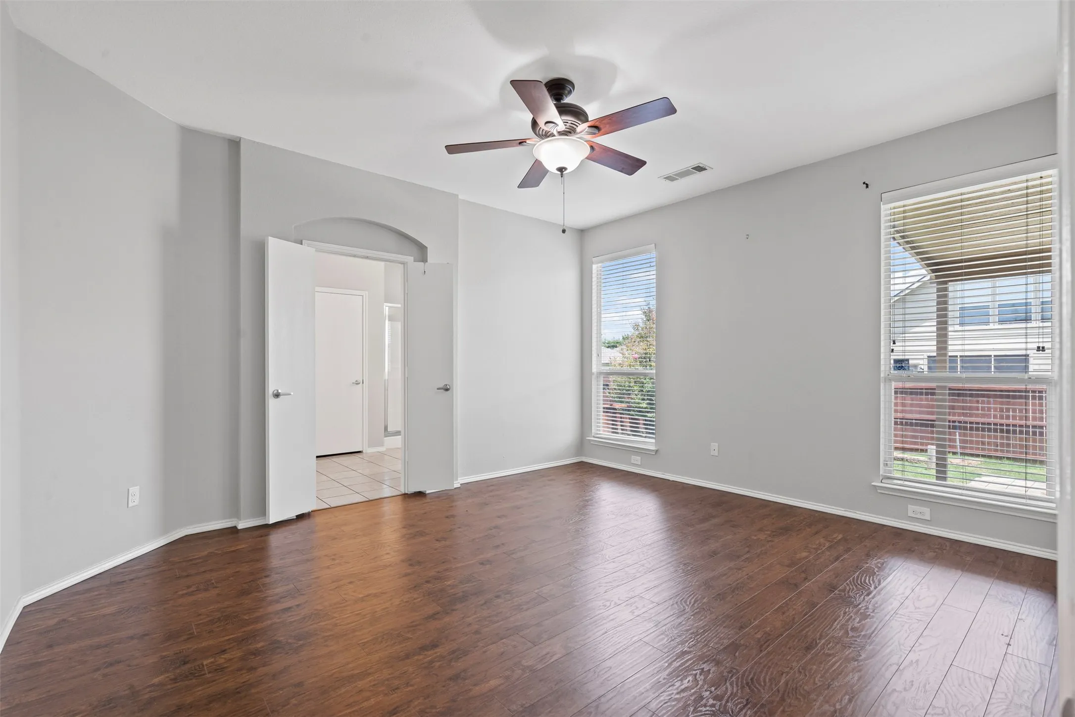 Wood-finish flooring, ceiling fan with integrated lighting, two windows with blinds, and double doors opening to an arched doorway