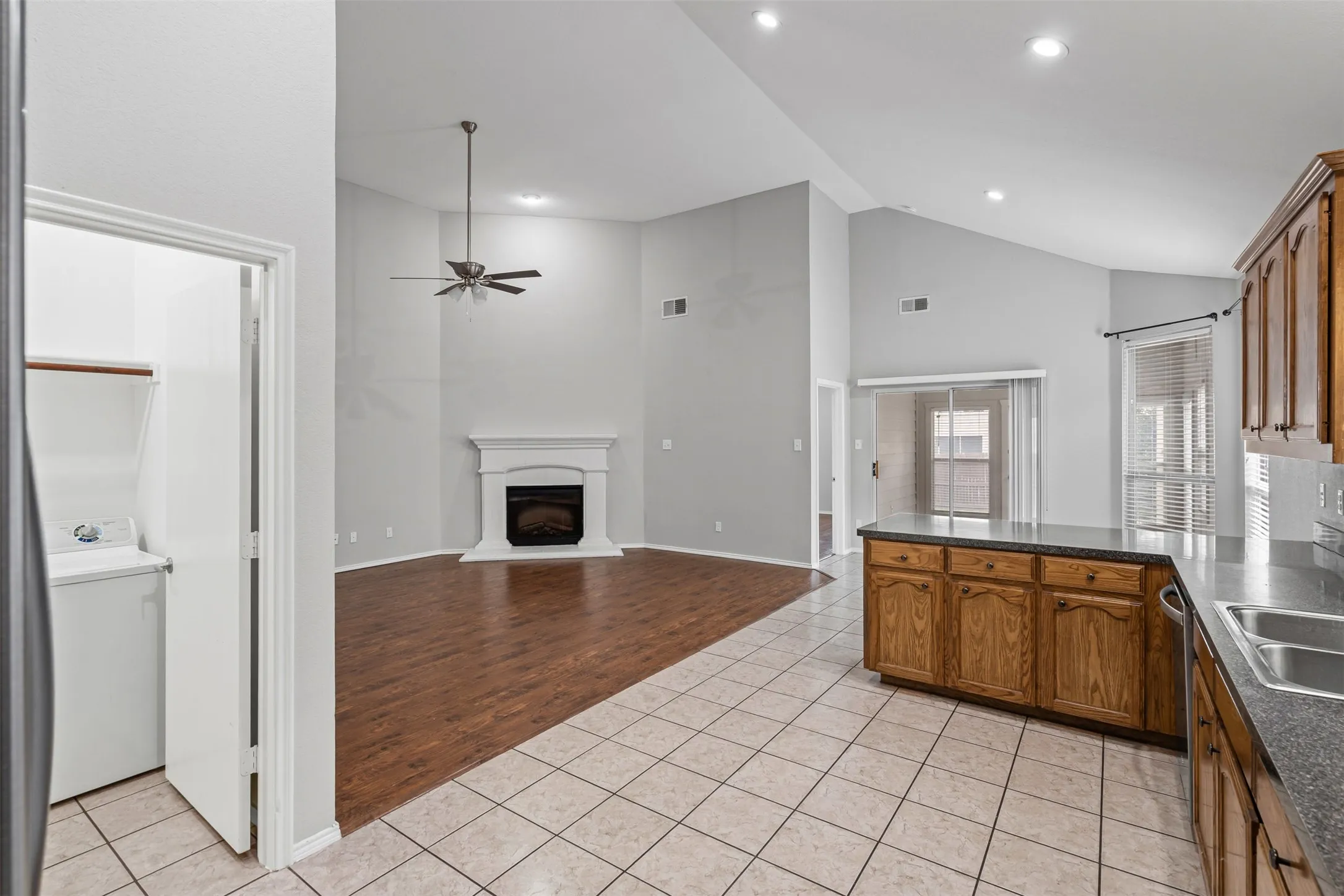 Vaulted ceiling with recessed lighting, wood-finish flooring, white fireplace with mantel, kitchen island with built-in cabinetry and countertop, and a double basin stainless steel sink