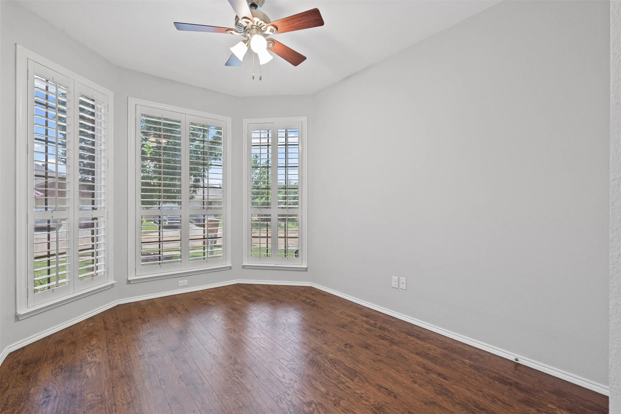Bay window with plantation shutters, wood-finish flooring, ceiling fan, and light gray wall finish