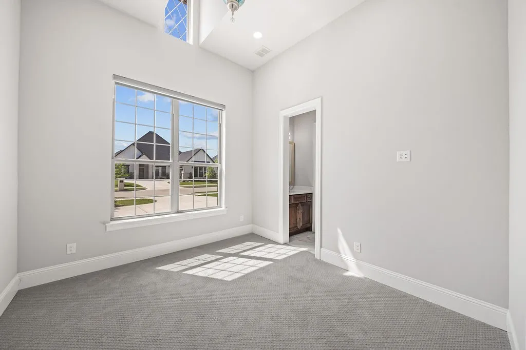 High ceilings with an upper arched window, a large rectangular window with grid panes, plush grey carpeting, and a doorway opening to a vanity with wood-finish cabinetry