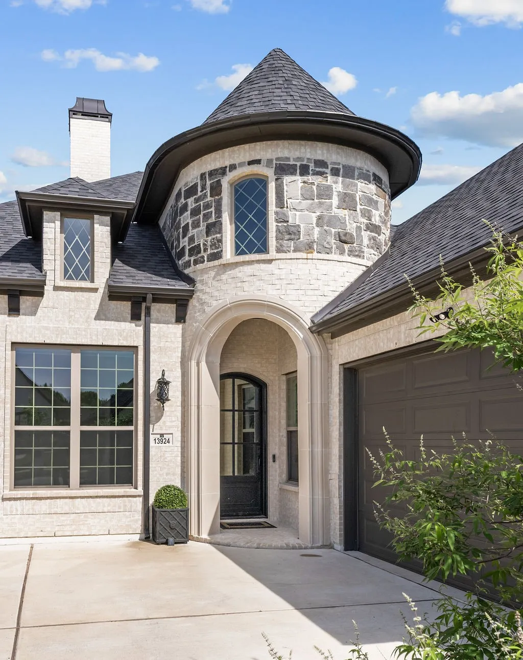 Turreted entry with an arched doorway, stone and brick facade, a dark asphalt shingle roof, and an integrated garage