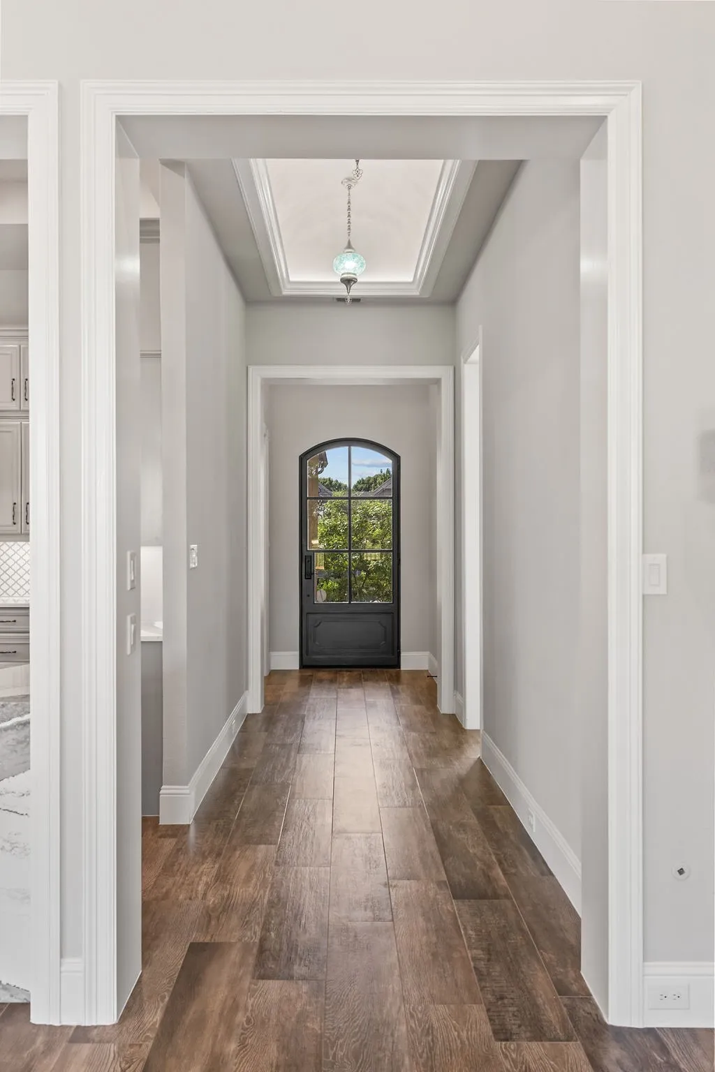 Wood-finish flooring, recessed ceiling with a decorative pendant light, arched entryway with a glass-paned door