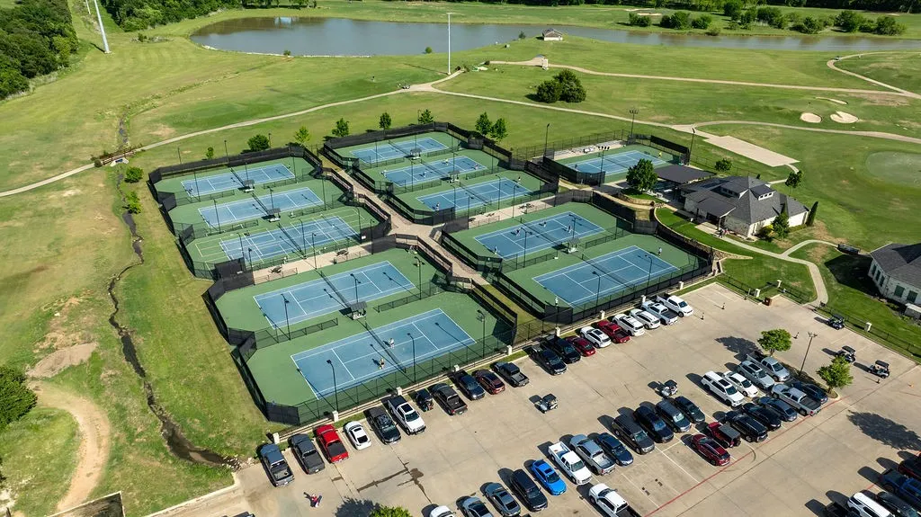 Multiple tennis courts with blue playing surfaces and green surrounds, adjacent to a large parking area
