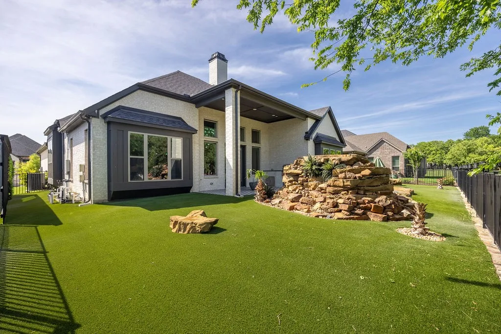 Expansive outdoor space featuring artificial turf, a rock water feature, dark privacy fencing, and a covered patio with columns