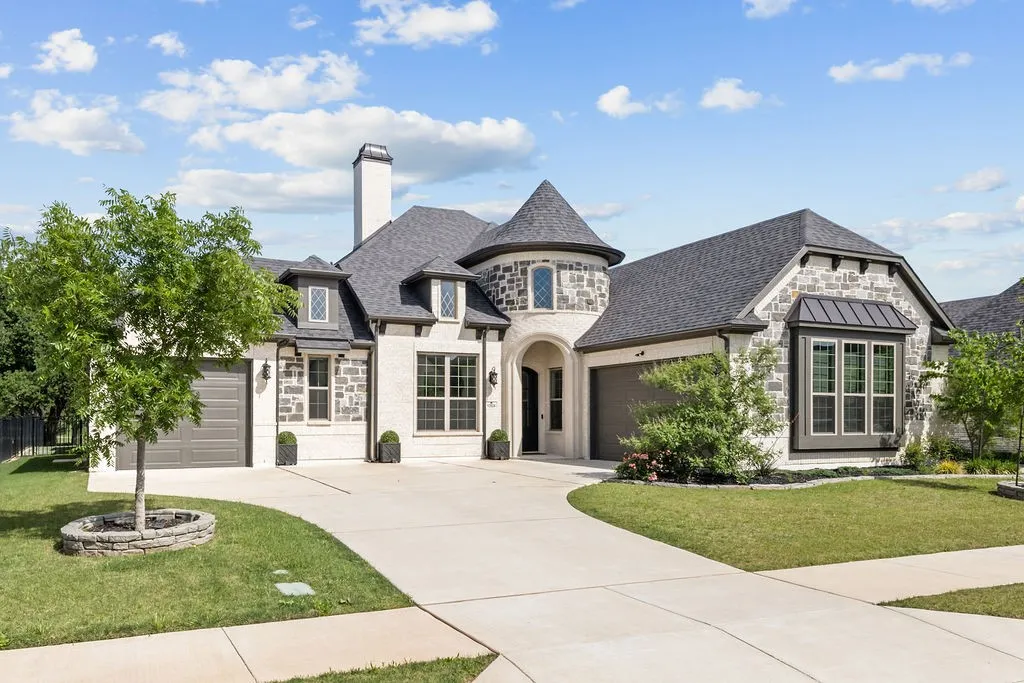 Stone and stucco exterior, prominent turret, dark gray garage doors, bay window with metal awning, and a curved concrete driveway