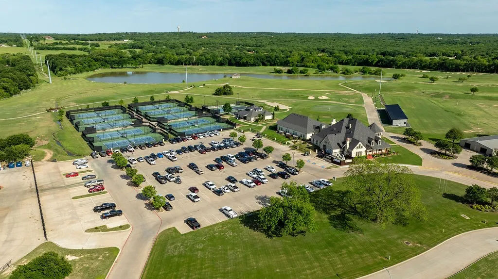 Expansive property with a large clubhouse, multiple tennis courts, a surface parking lot, and a natural pond feature, all surrounded by green space
