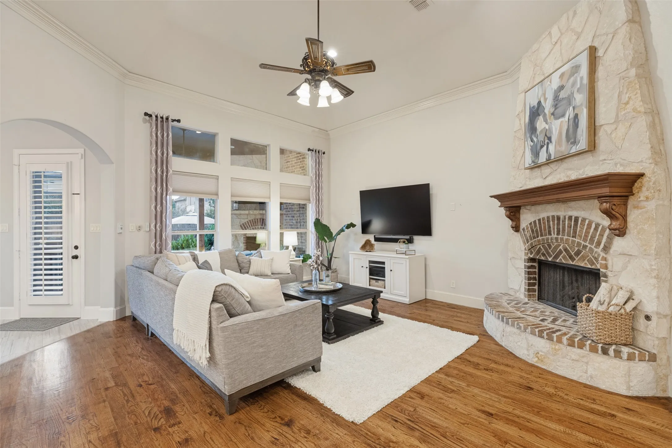 Vaulted ceiling with crown molding, stone-clad fireplace with brick accents, arched doorways, wood-finish flooring, and a ceiling fan with integrated lighting