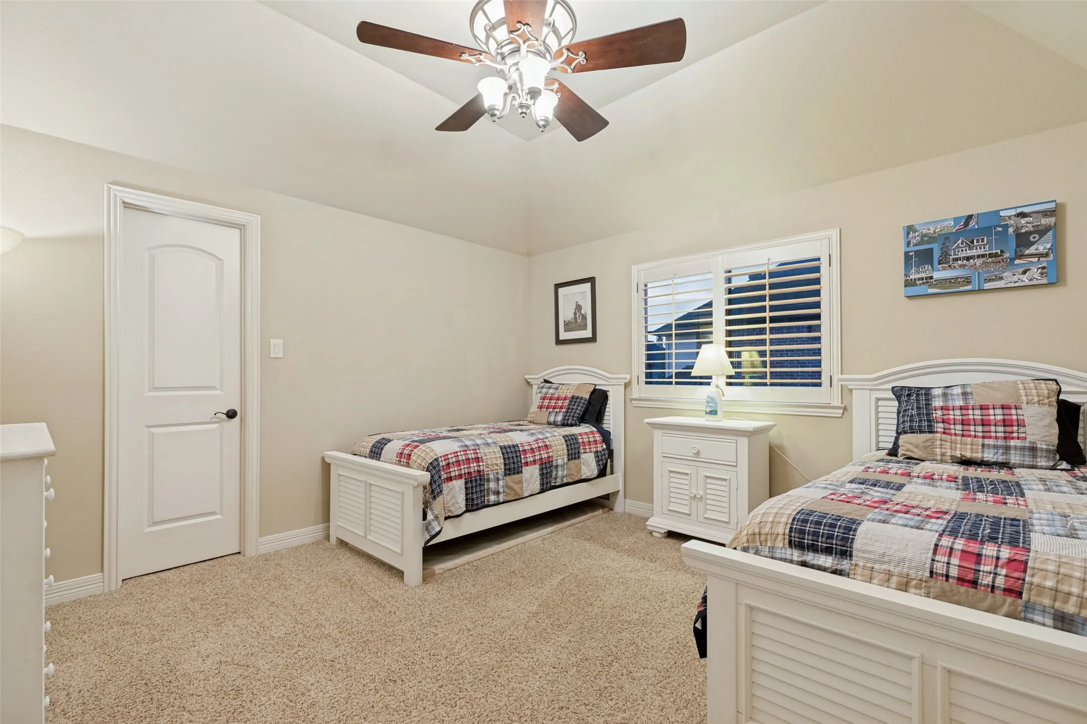 Vaulted ceiling, ceiling fan with integrated lighting, window with white plantation shutters, neutral tone wall paint, and light-colored carpet flooring