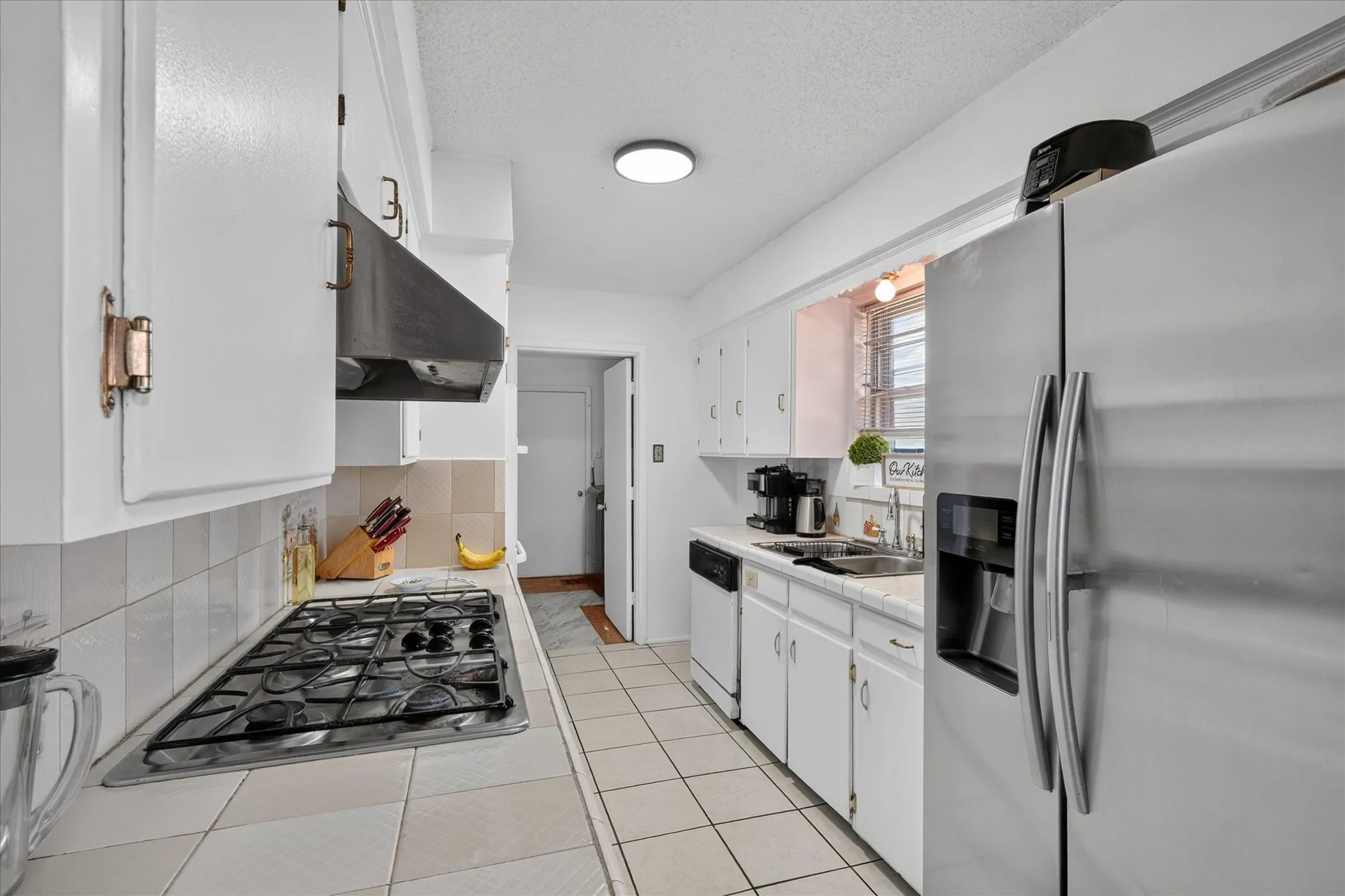Galley kitchen featuring white cabinetry, a gas cooktop with an overhead exhaust hood, a stainless steel refrigerator, and tiled flooring