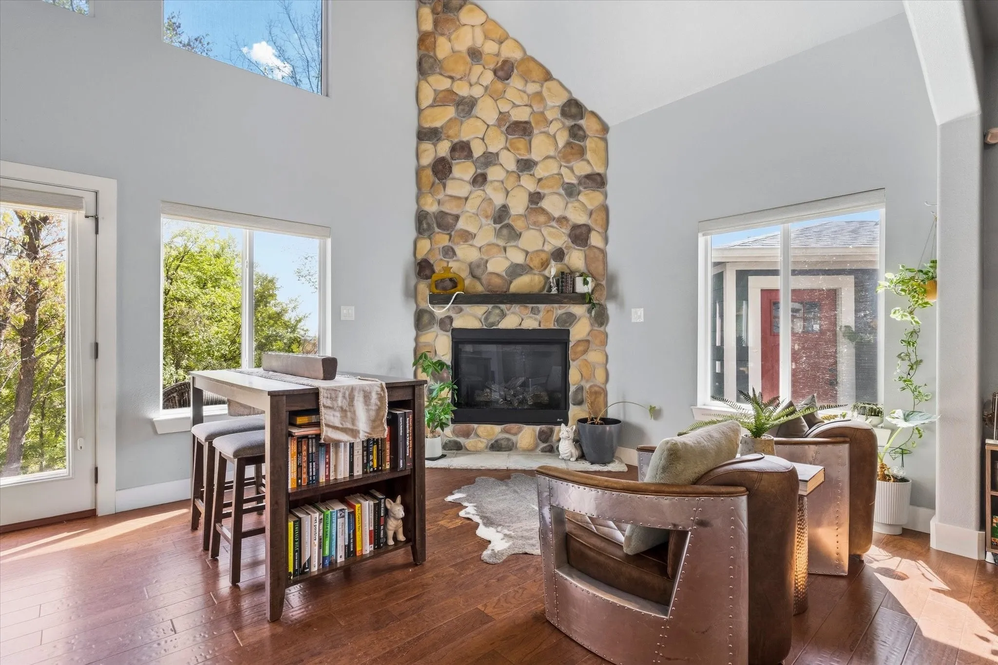 Soaring vaulted ceilings, a prominent stone-clad fireplace with a wood mantel, rich wood-finish flooring, and multiple large windows