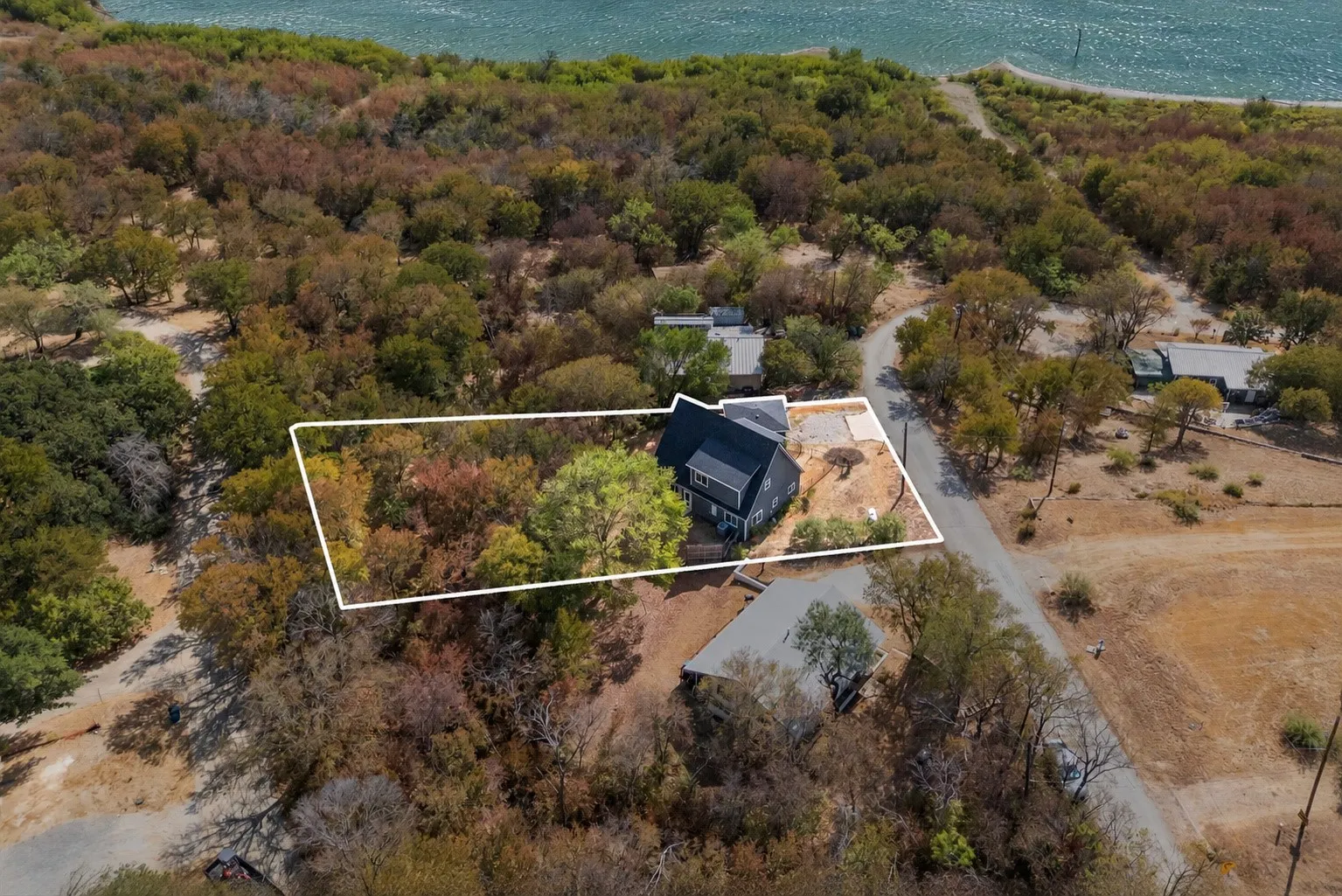 Aerial property view featuring a dark-toned roofline, surrounding mature trees, and a visible water body