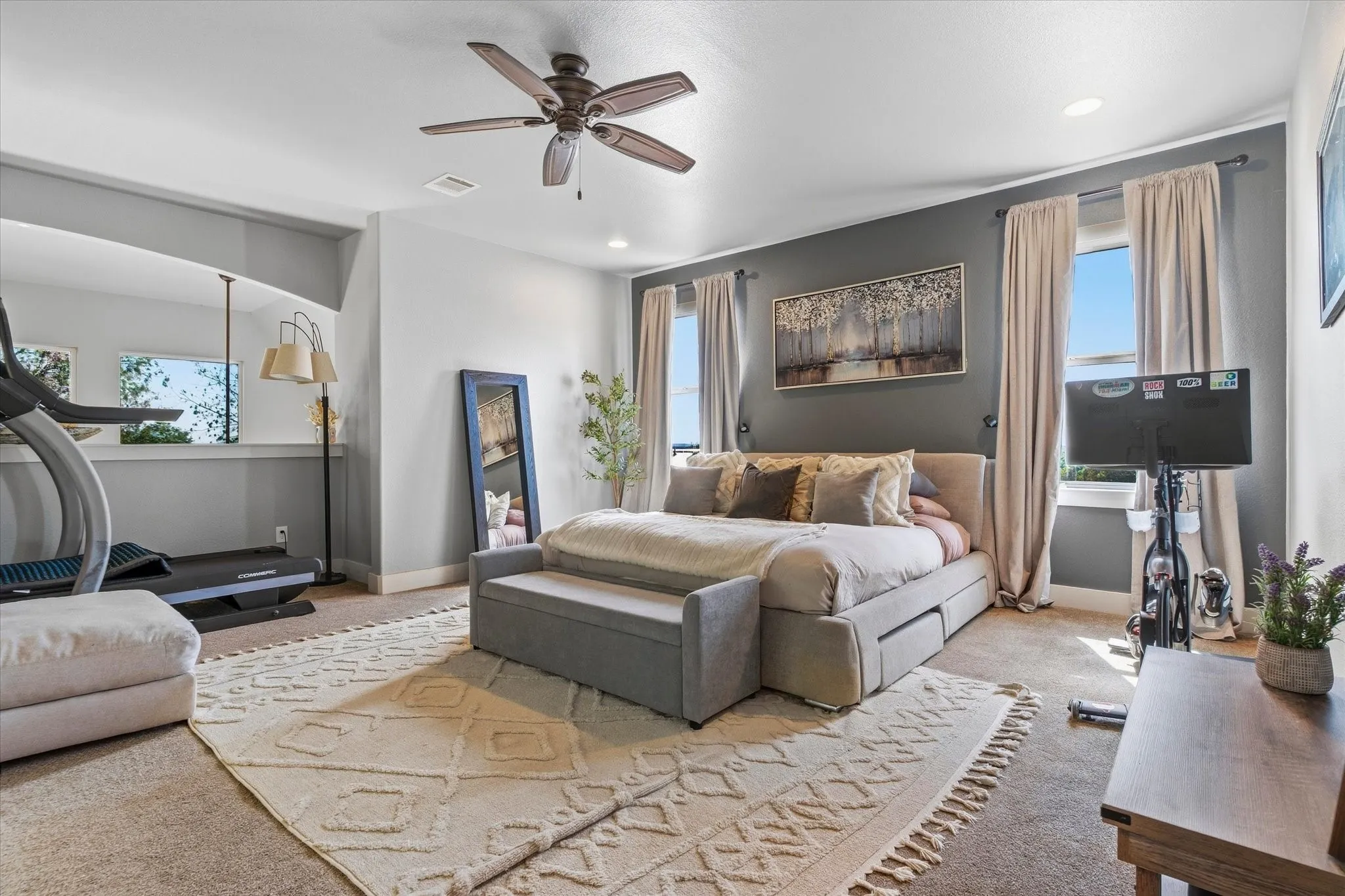 Spacious primary bedroom featuring ceiling fan with light fixture, wood-finish flooring, layered area rugs, and multiple windows with thermal curtains