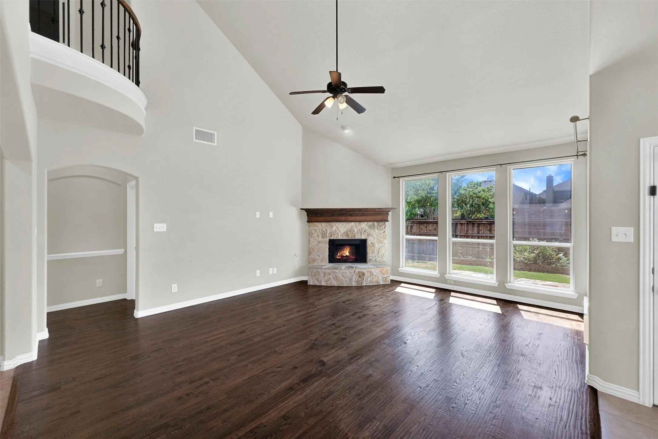 Vaulted ceiling with a ceiling fan, stone-faced fireplace with a wood mantle, wood-finish flooring, and multiple large windows