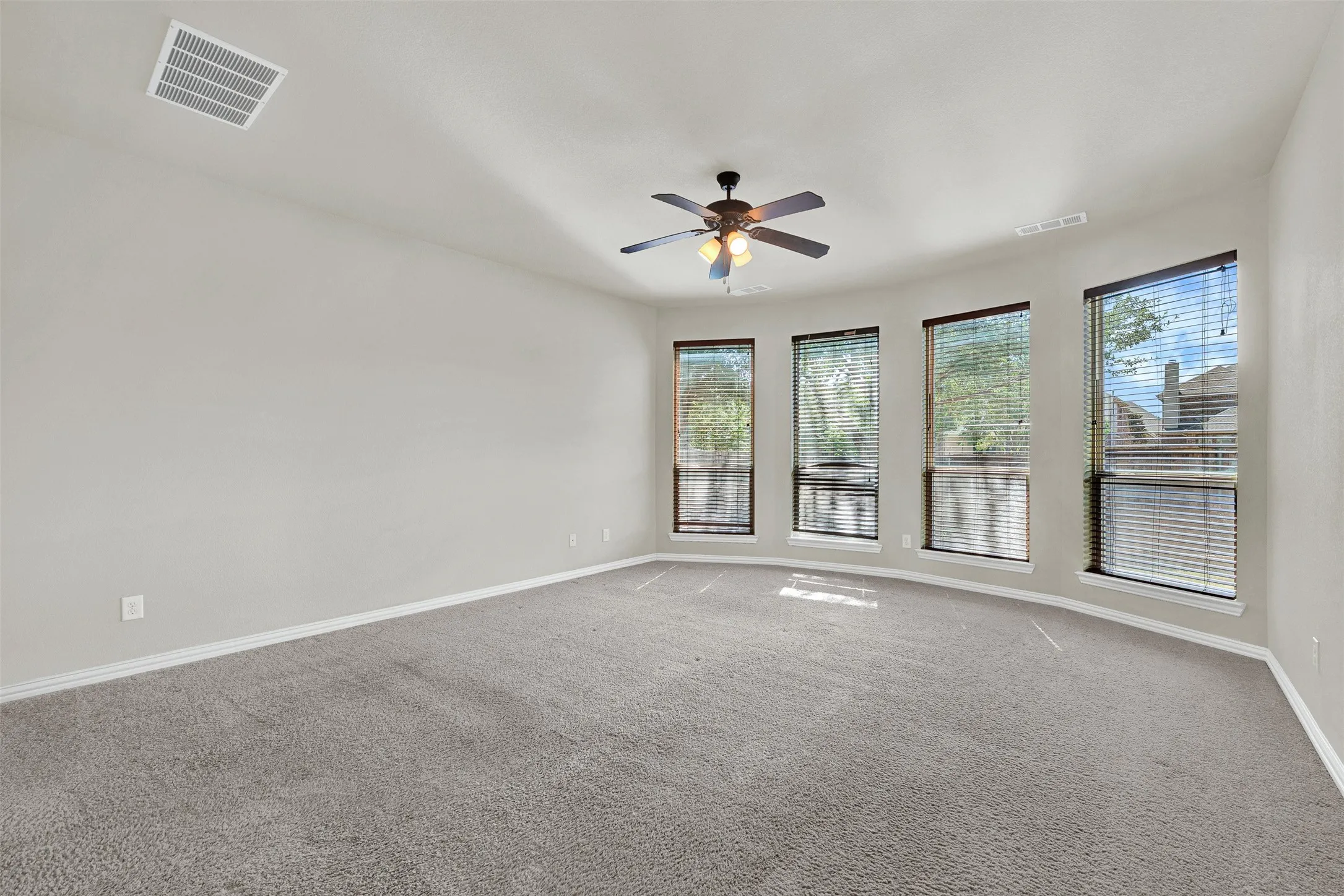 Spacious room featuring curved wall, four windows with blinds, gray carpet, and a ceiling fan with integrated lighting