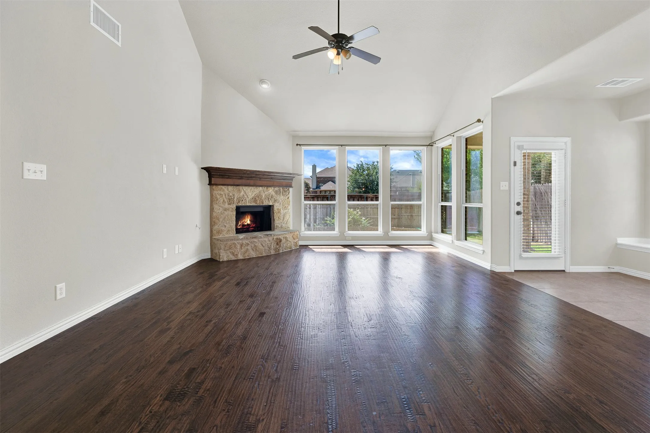 Vaulted ceilings with a ceiling fan, stone-surround fireplace with wood mantle, dark wood-finish flooring, and a wall of windows