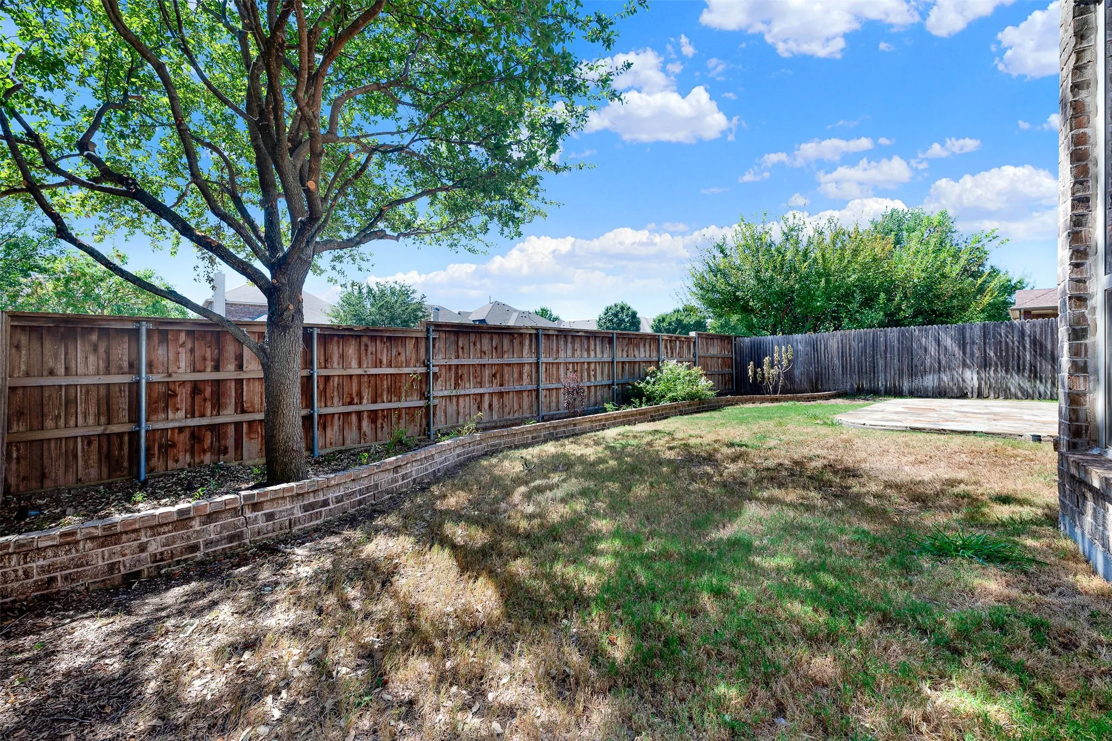 Expansive rear yard featuring mature trees, wood privacy fencing, a brick retaining wall, and a concrete patio area