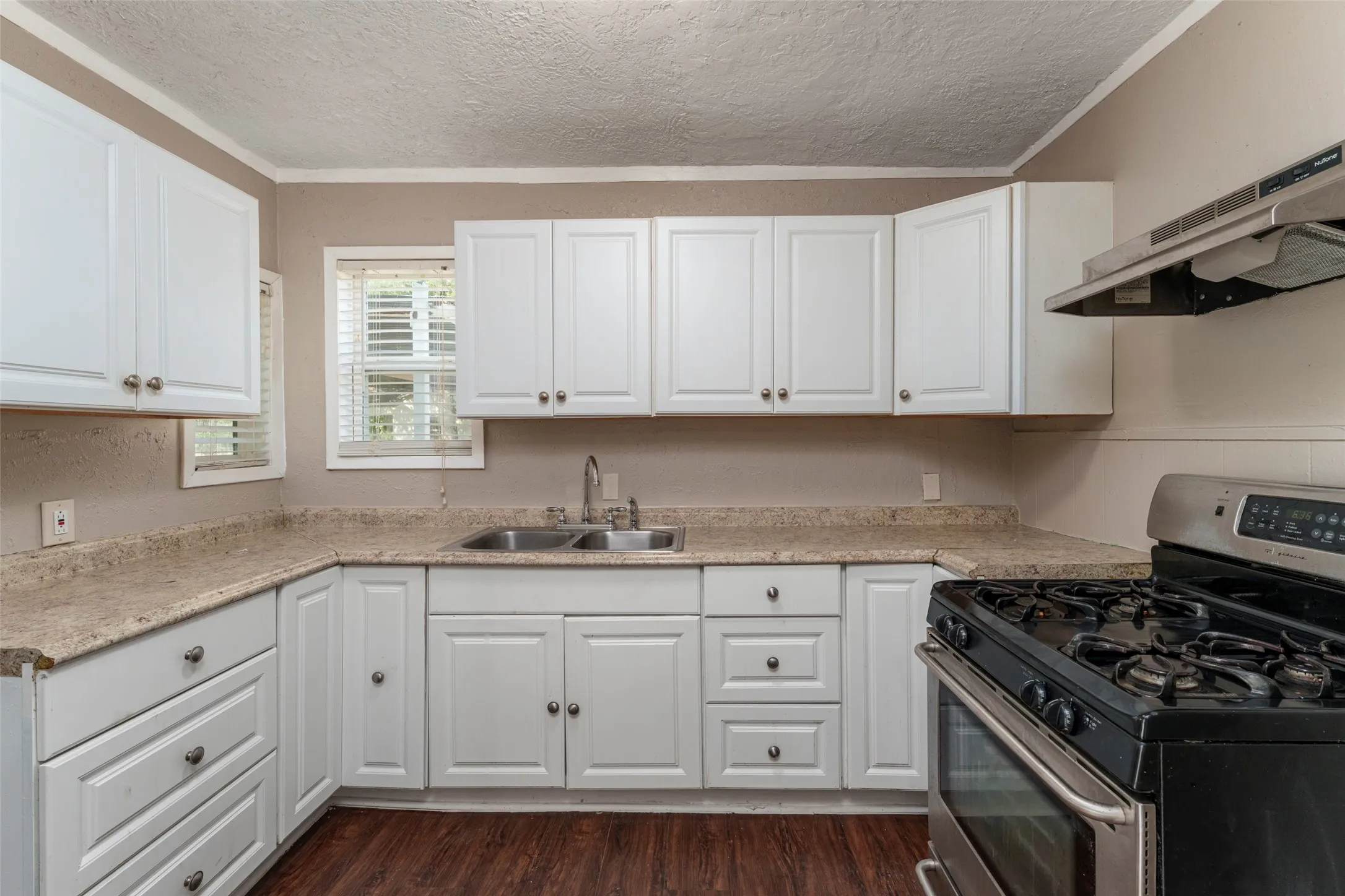 White cabinetry with silver hardware, dual basin stainless steel sink, light-toned countertops, range with stainless steel hood, and wood-finish flooring