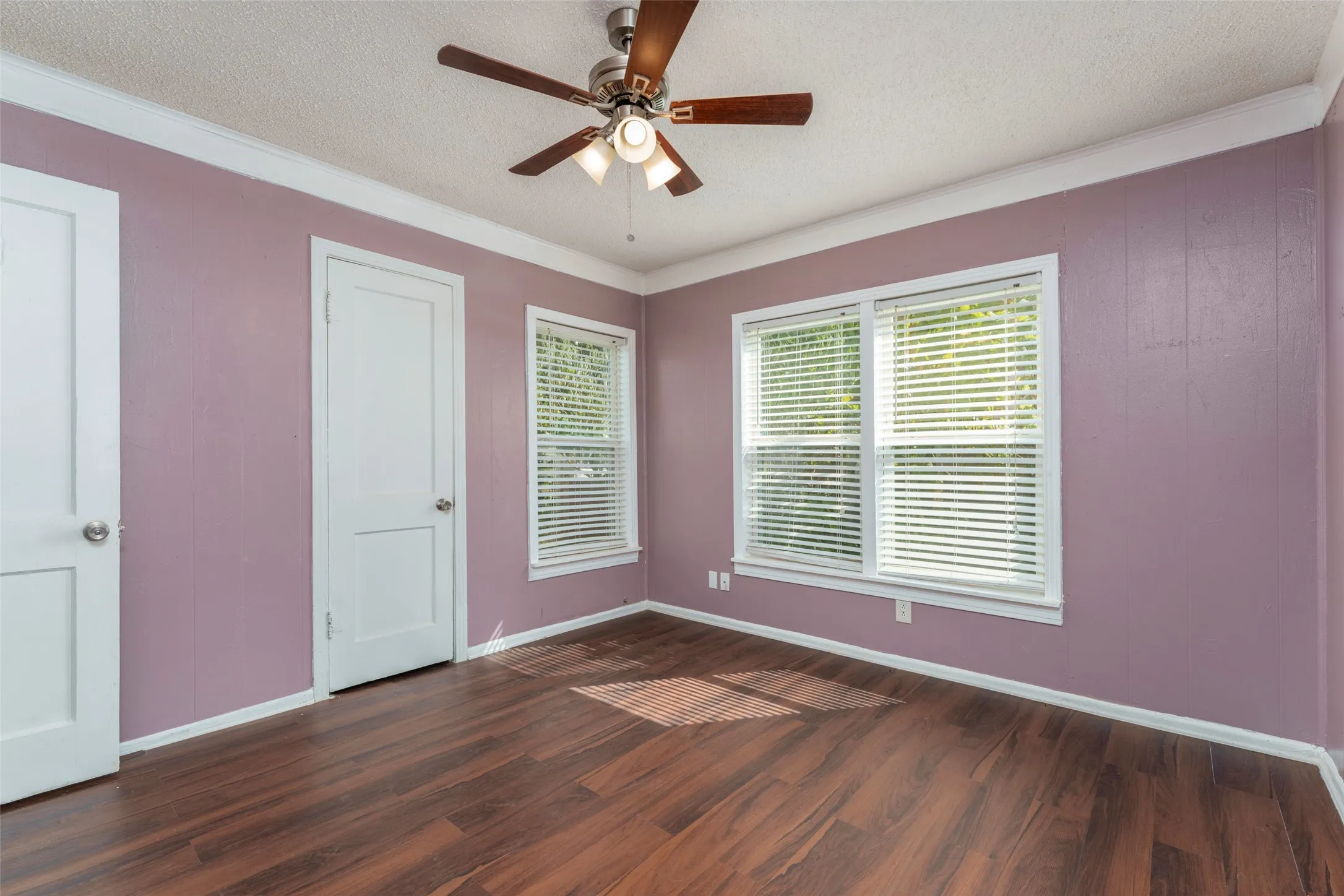 Wood-finish flooring, white trim, dual windows with blinds, and a ceiling fan with integrated lighting