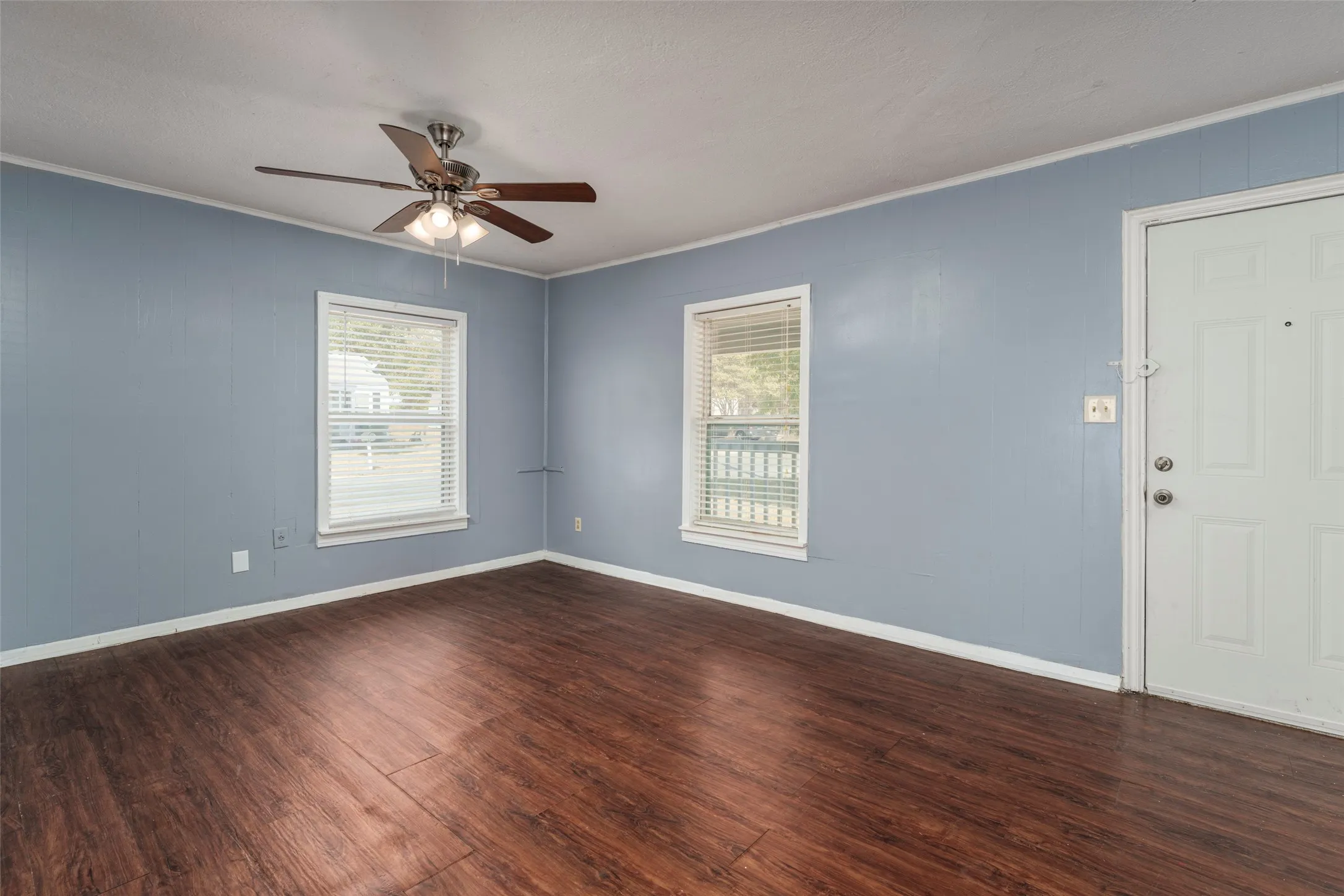 Wood-finish flooring throughout, two windows with blinds, ceiling fan with integrated lighting, and a paneled entry door