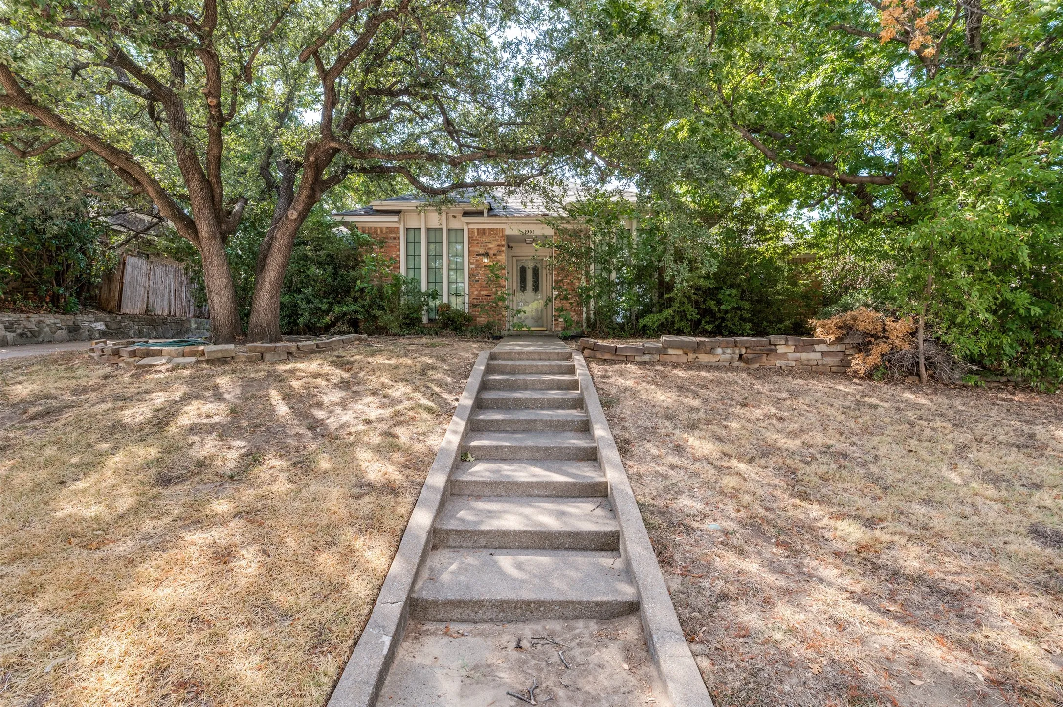Brick facade residence with a concrete walkway, mature trees, and a stone retaining wall