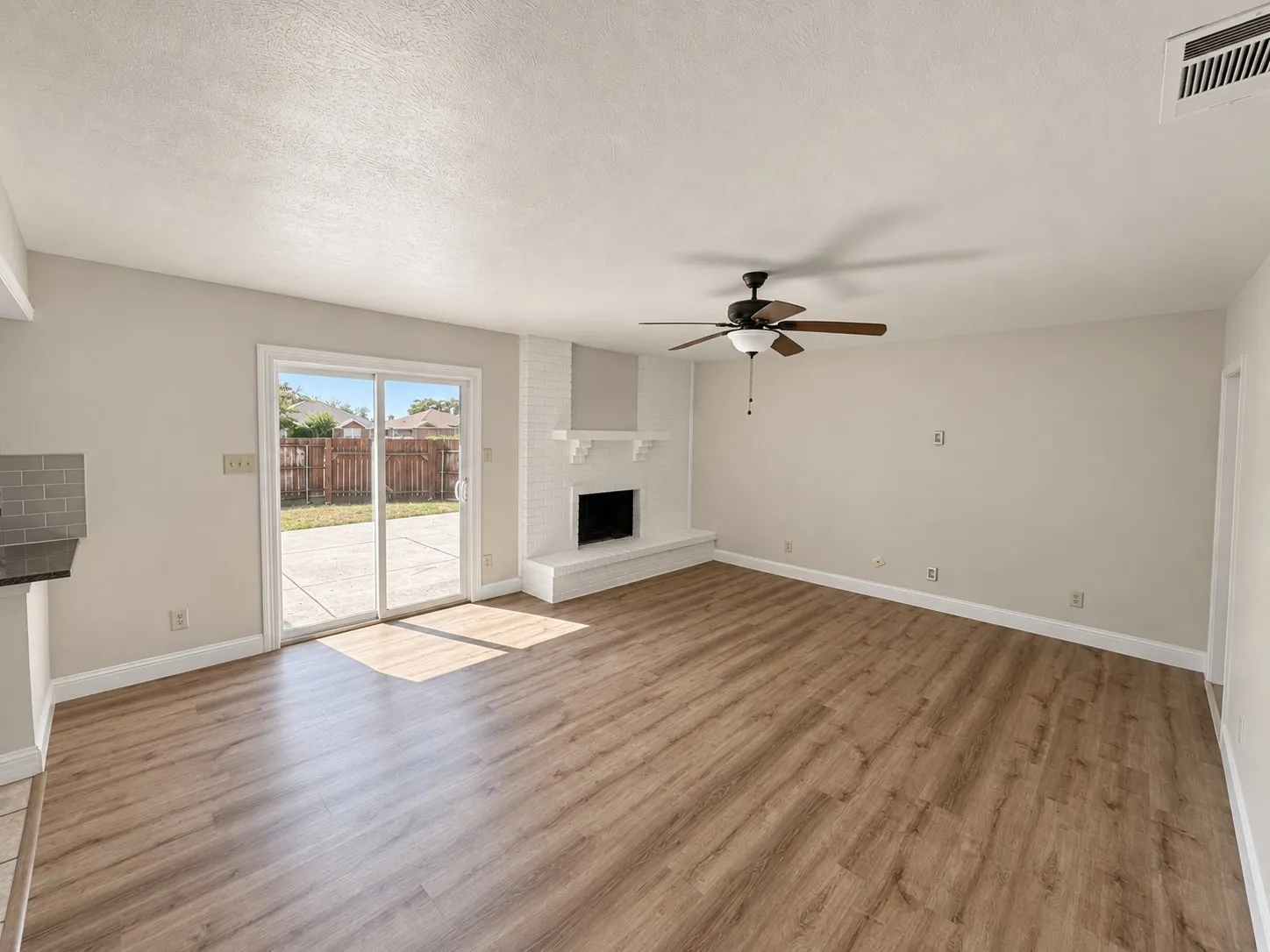 Wood-finish flooring throughout, sliding glass door leading to a concrete patio, white brick fireplace with mantel, ceiling fan with integrated lighting, and gray subway tile backsplash