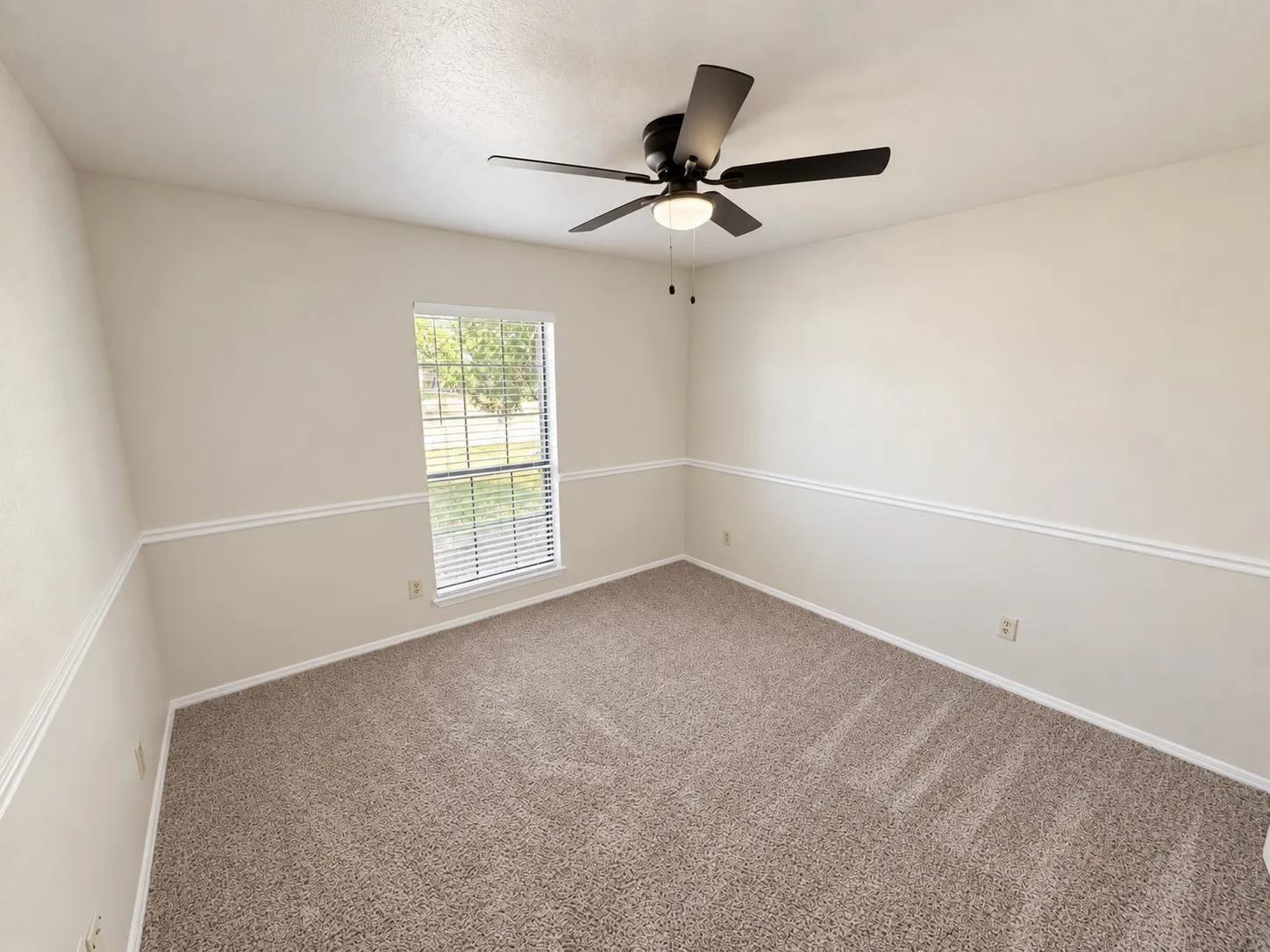 Neutral wall paint with chair rail molding, textured carpet flooring, window with blinds, and a ceiling fan with integrated lighting