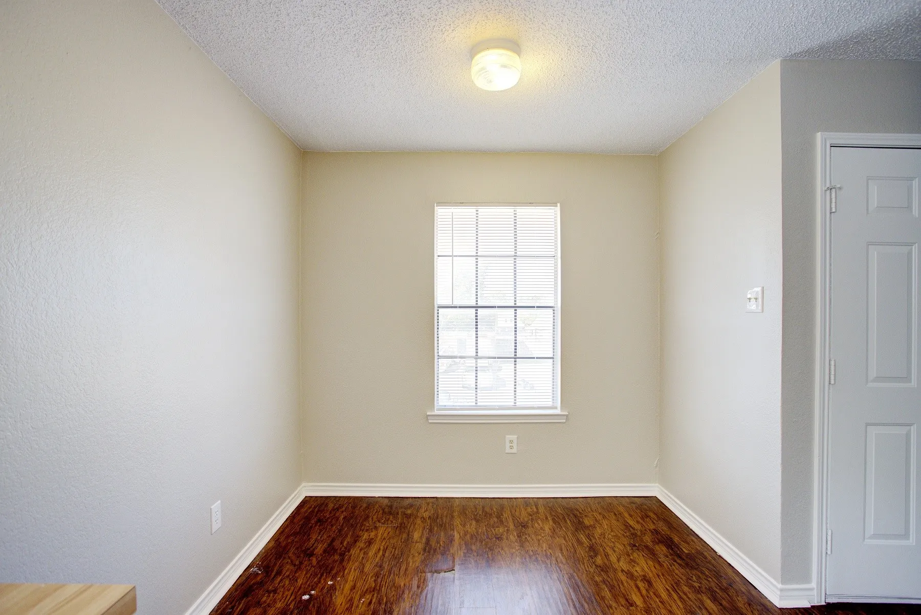 Wood-finish flooring with white trim, a single window with blinds, and an overhead flush-mount light fixture