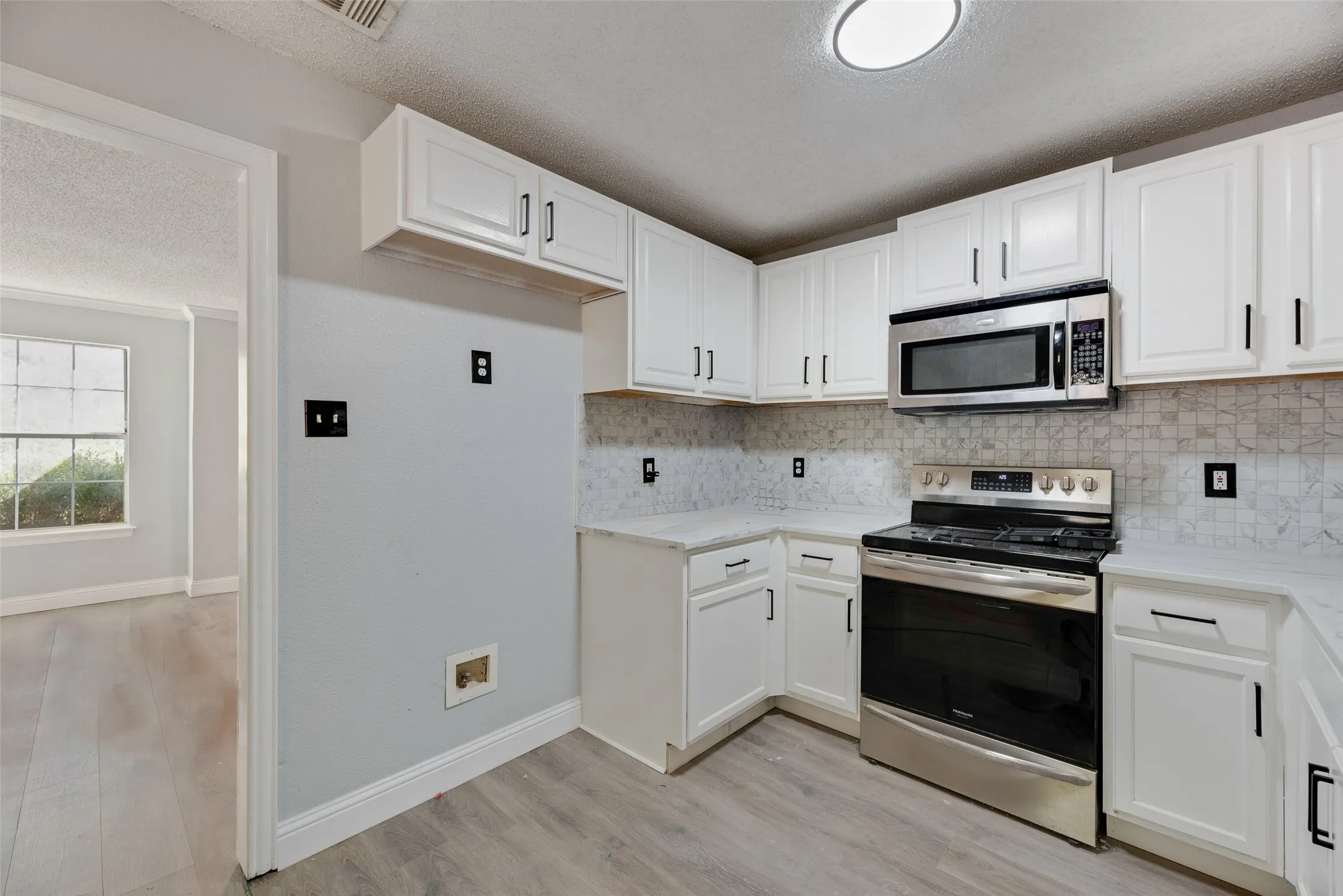 White cabinetry with black hardware, stainless steel appliances, patterned backsplash, light-toned countertops, and wood-finish flooring