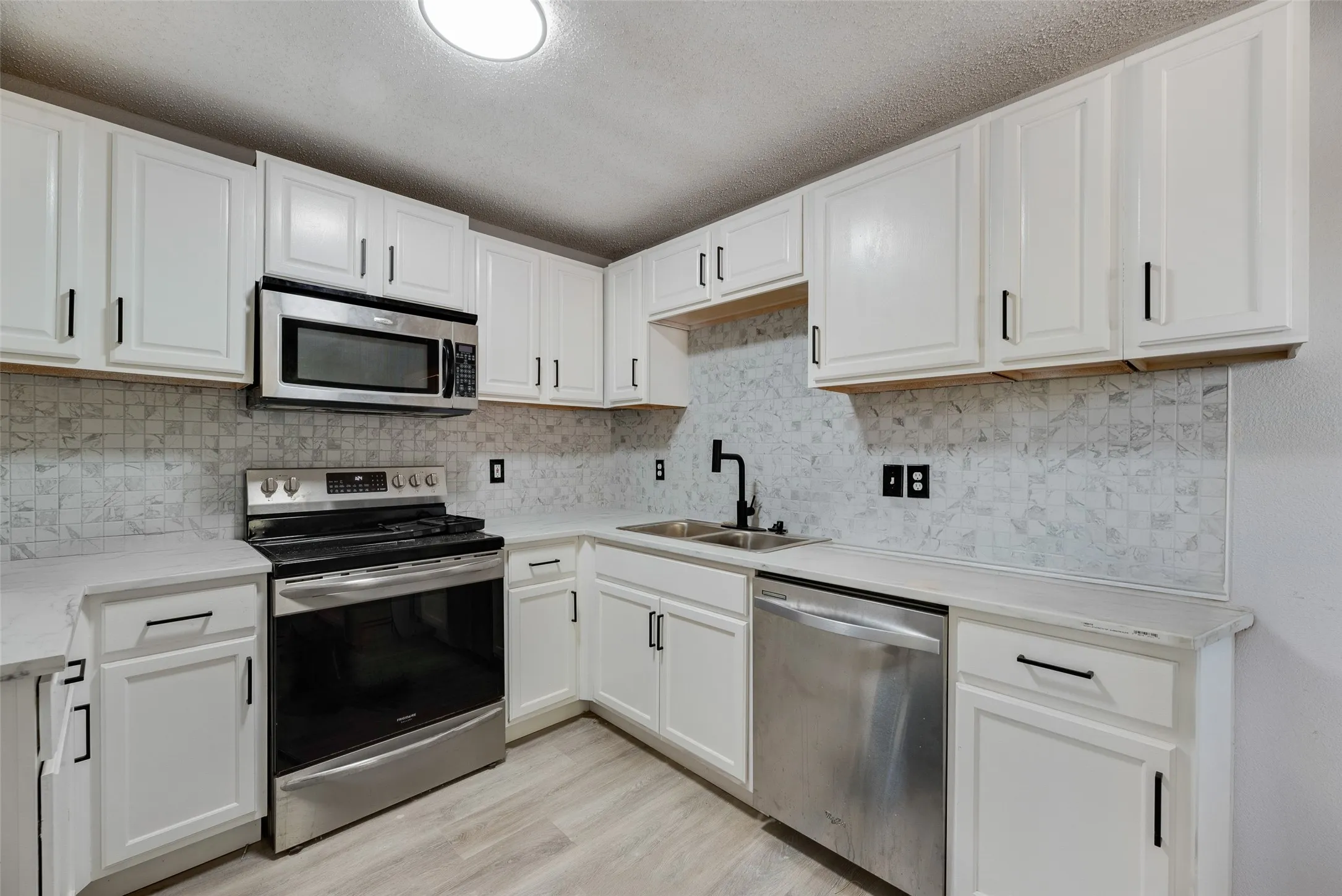 White cabinetry with matte black hardware, stainless steel appliances, patterned tile backsplash, light-toned countertops, and wood-finish flooring