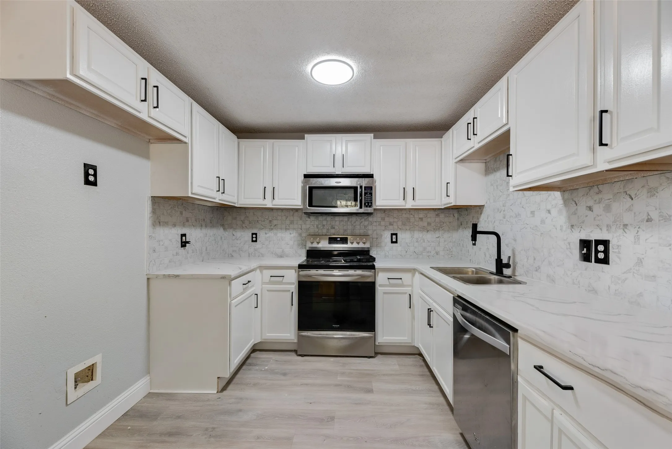White shaker-style cabinetry with black hardware, stainless steel appliances, light-toned countertops, and wood-finish flooring