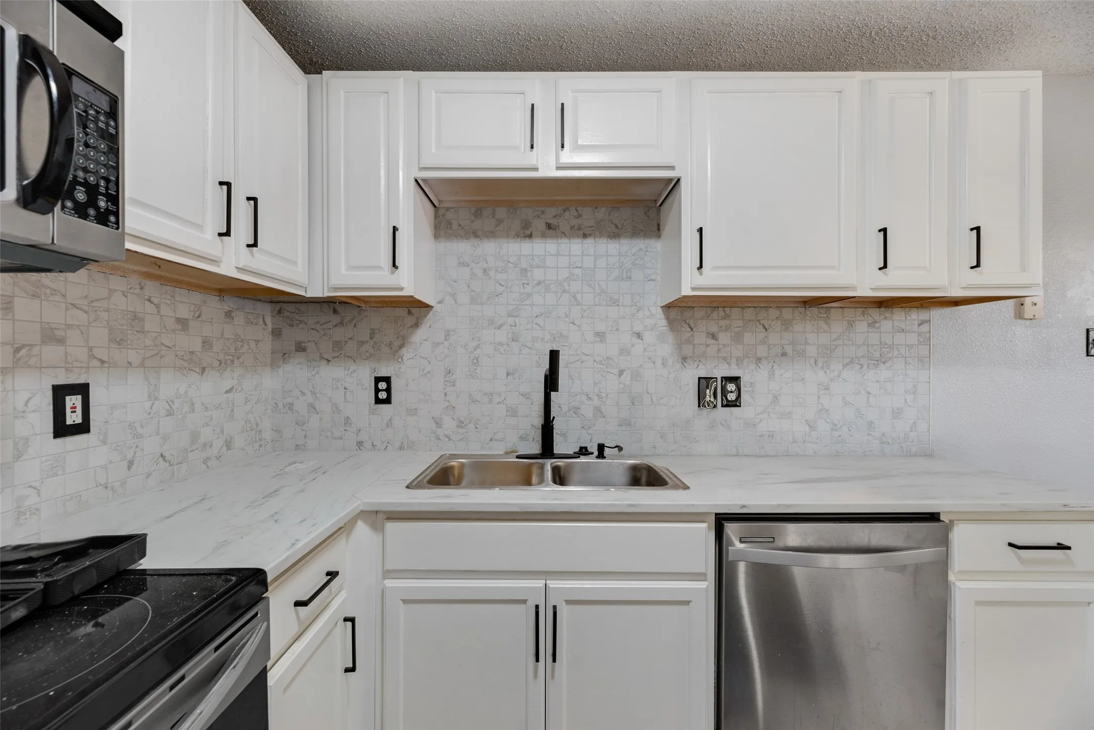 White shaker-style cabinetry with black hardware, stainless steel dishwasher, double-basin sink with a matte black faucet, and patterned tile backsplash
