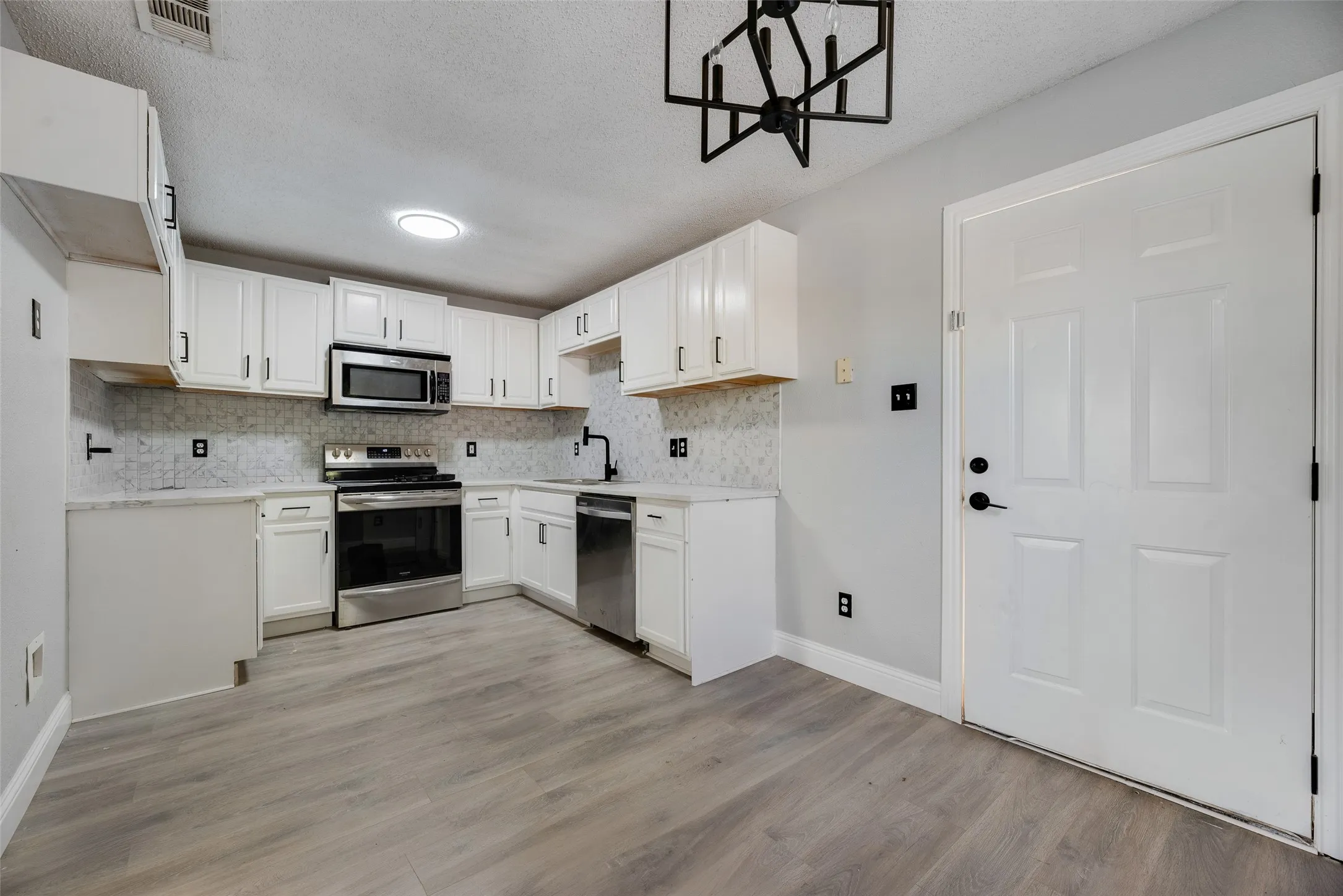 White cabinetry with matte black hardware, stainless steel appliances, patterned backsplash, light-toned wood-finish flooring, and a contemporary ceiling fixture