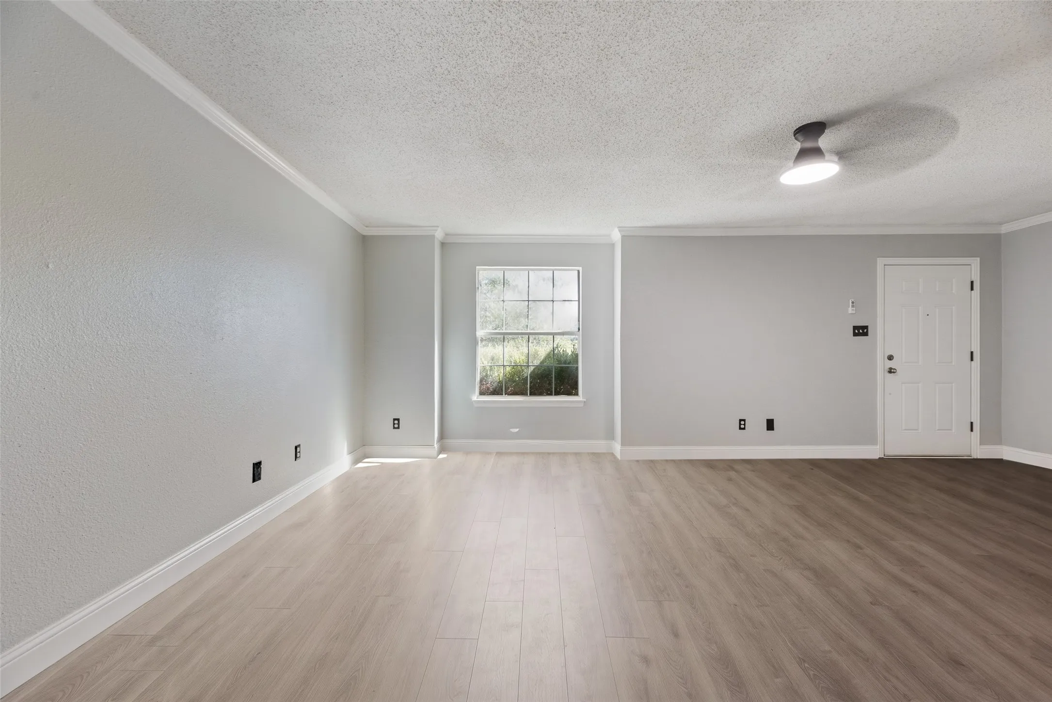 Expansive room featuring wood-finish flooring, neutral-toned walls, a large window, a decorative ceiling light fixture, and a solid entry door