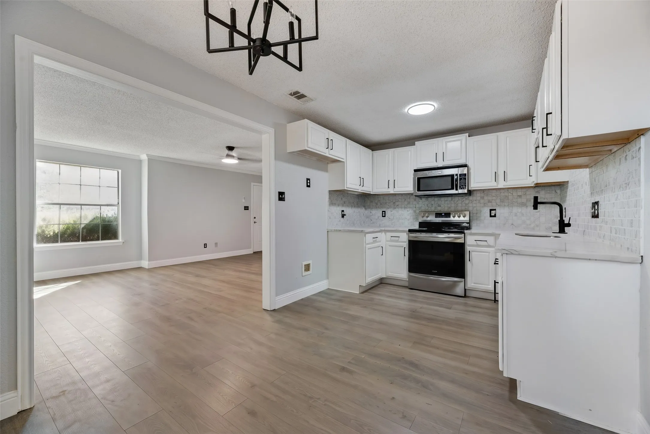 Light wood-finish flooring throughout, bright white kitchen cabinetry, stainless steel appliances, white tile backsplash, and a modern black linear chandelier