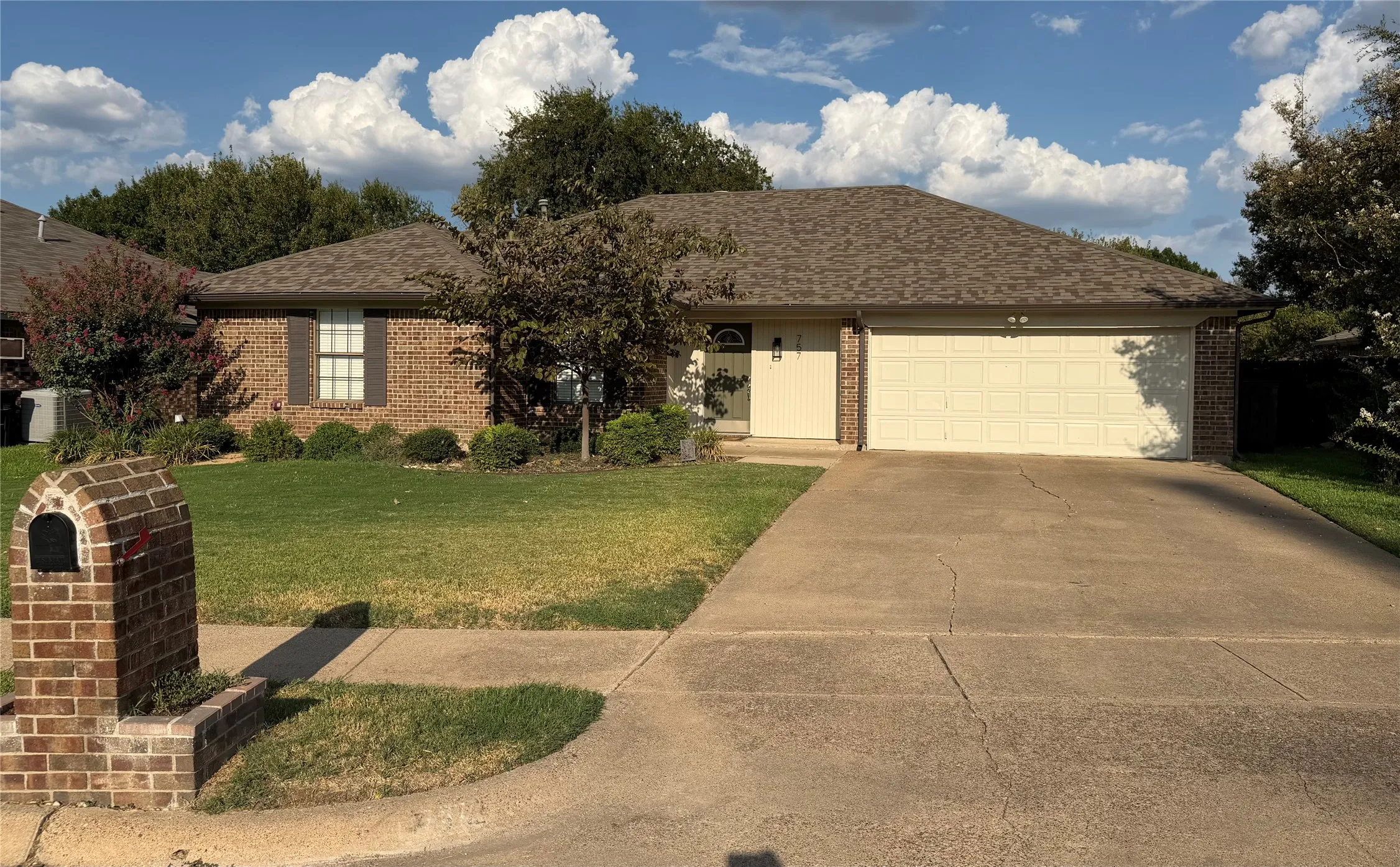 Brick exterior with brown shutters, attached two-car garage, concrete driveway, and a manicured lawn with mature landscaping