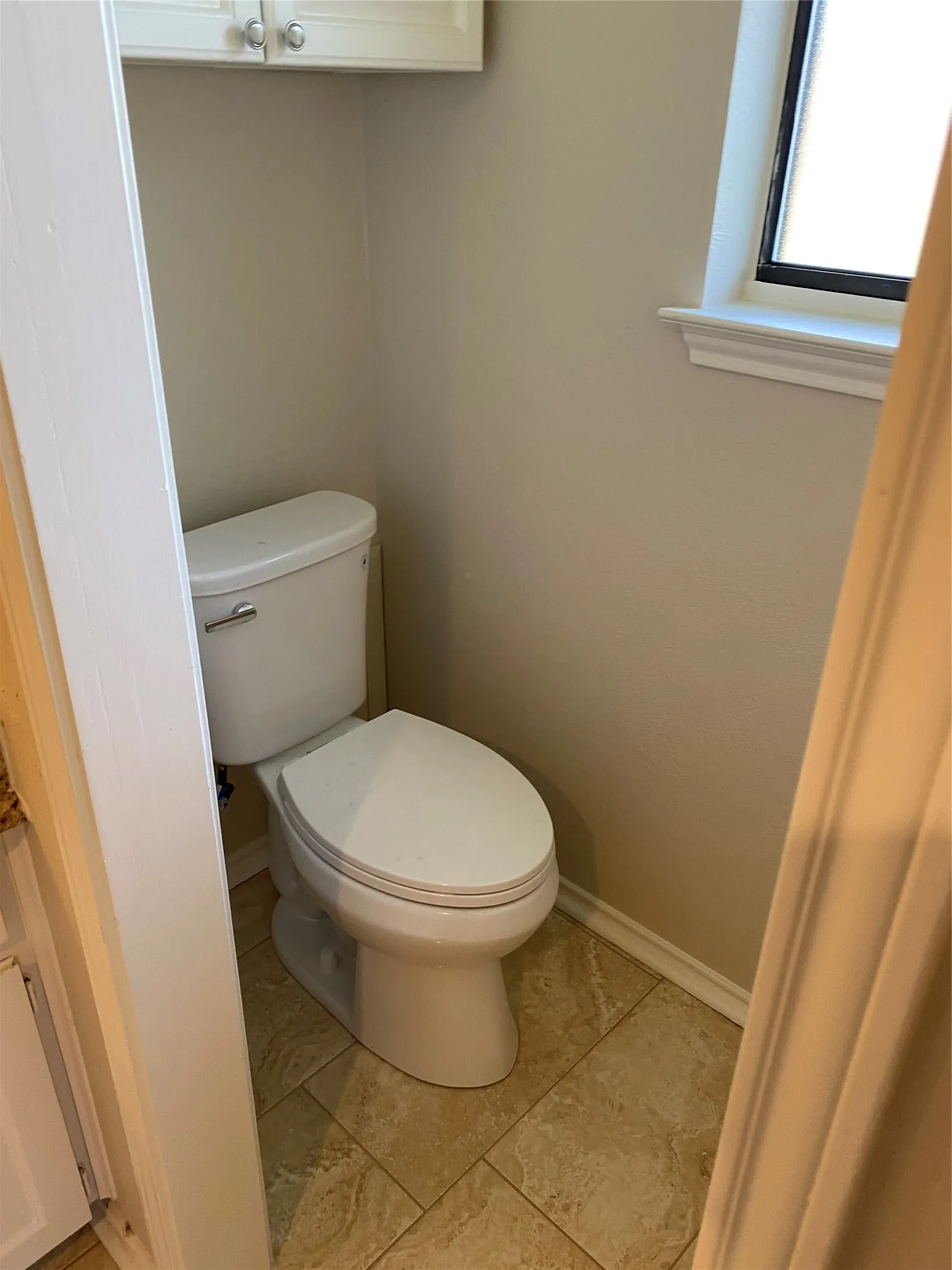 White toilet, tiled flooring, white cabinetry with silver-tone hardware, and a window with white trim